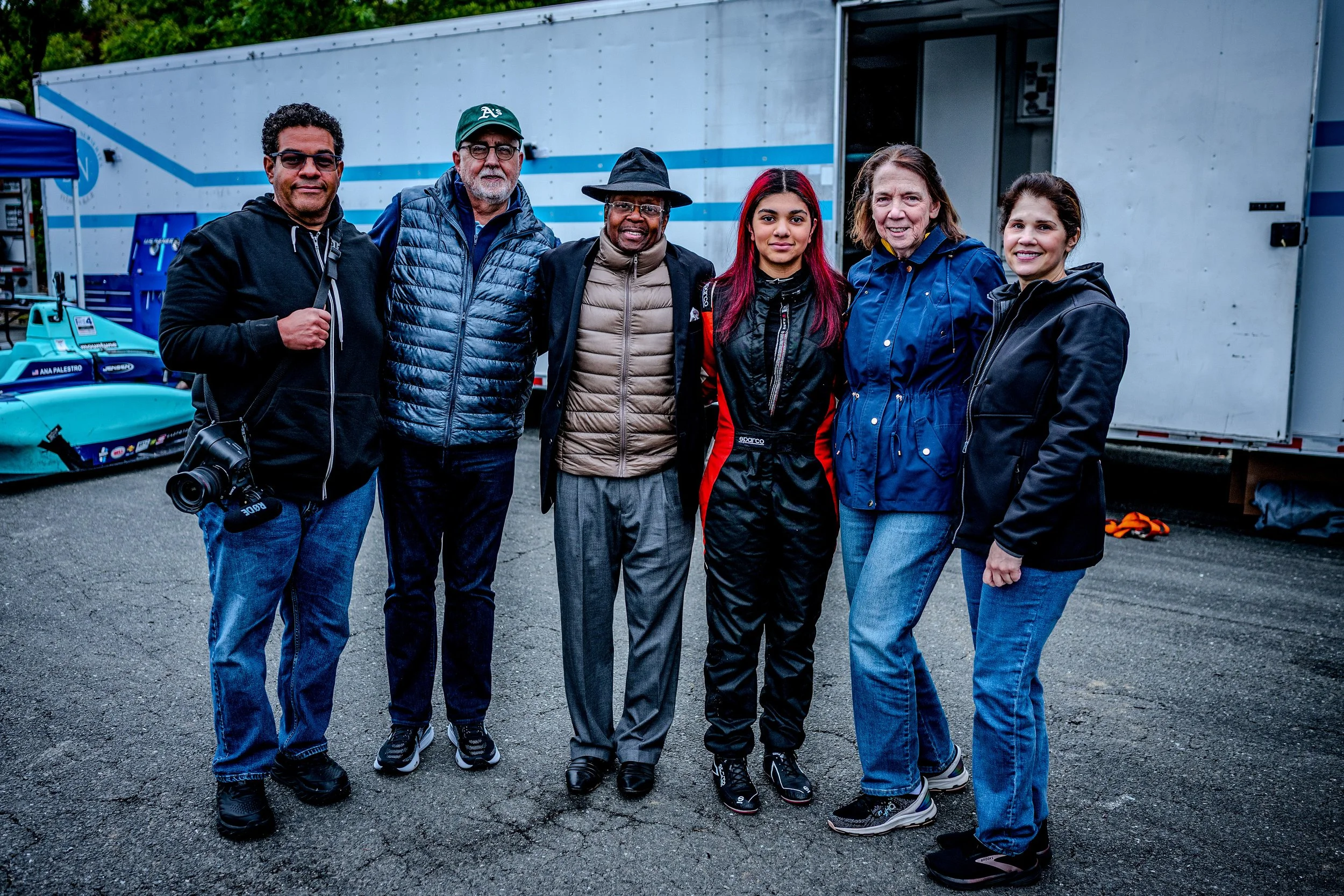 Group of seven diverse people standing together outdoors, smiling, with a trailer and racing car in the background.