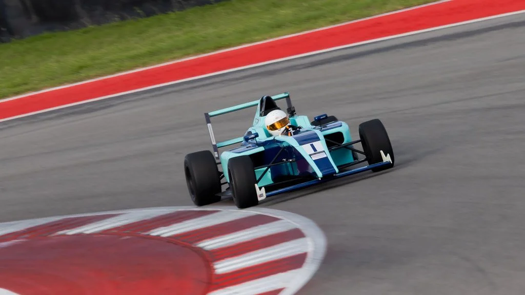 A blue and light blue open-wheel race car with the number 1 on the front, driving on a racing track and taking a turn at high speed.