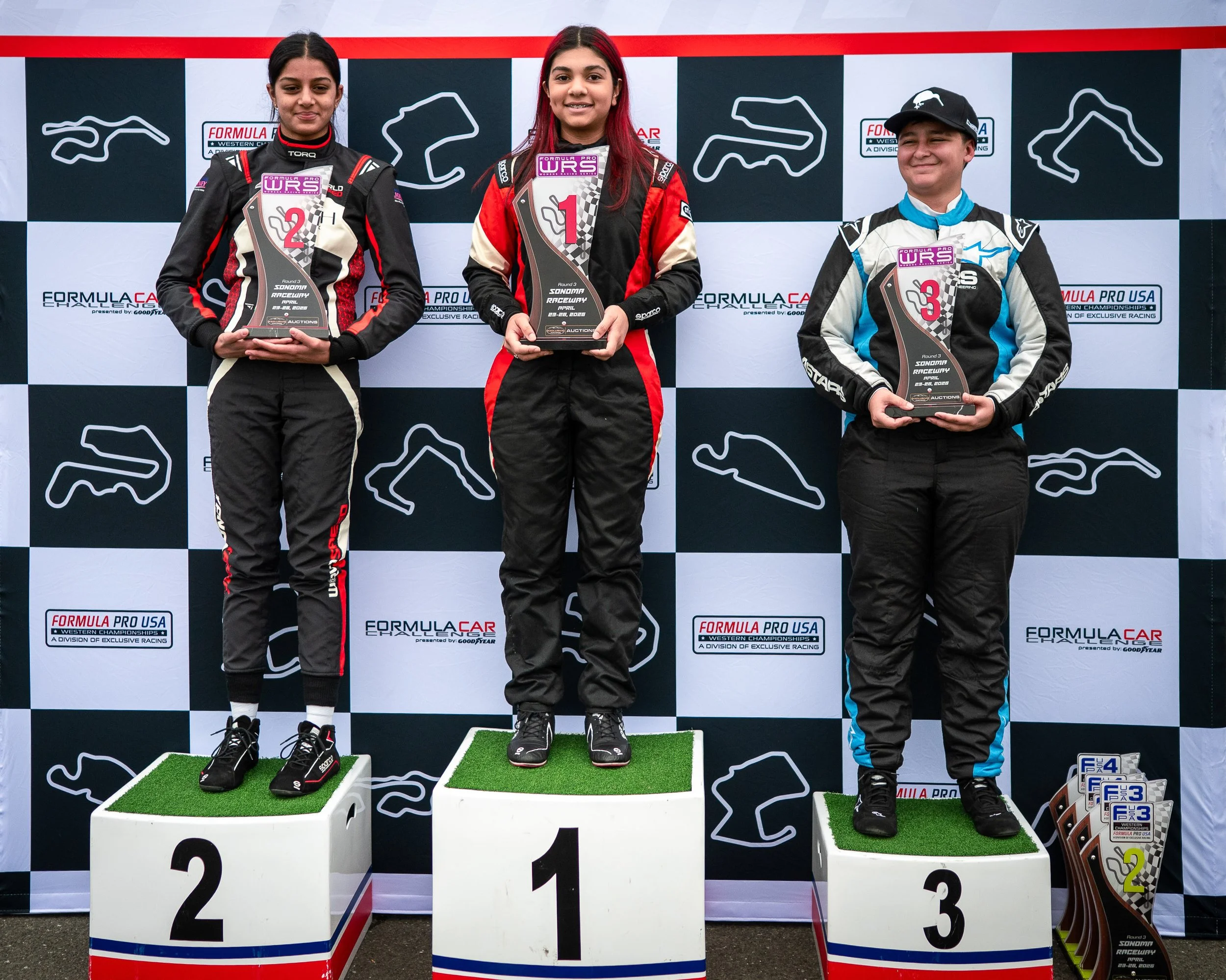 Three young race car drivers standing on winners' podiums holding trophies, with a checkered racing backdrop. The girl in the center is on the first place podium, the girl on the left is on the second place, and the boy on the right is on the third p