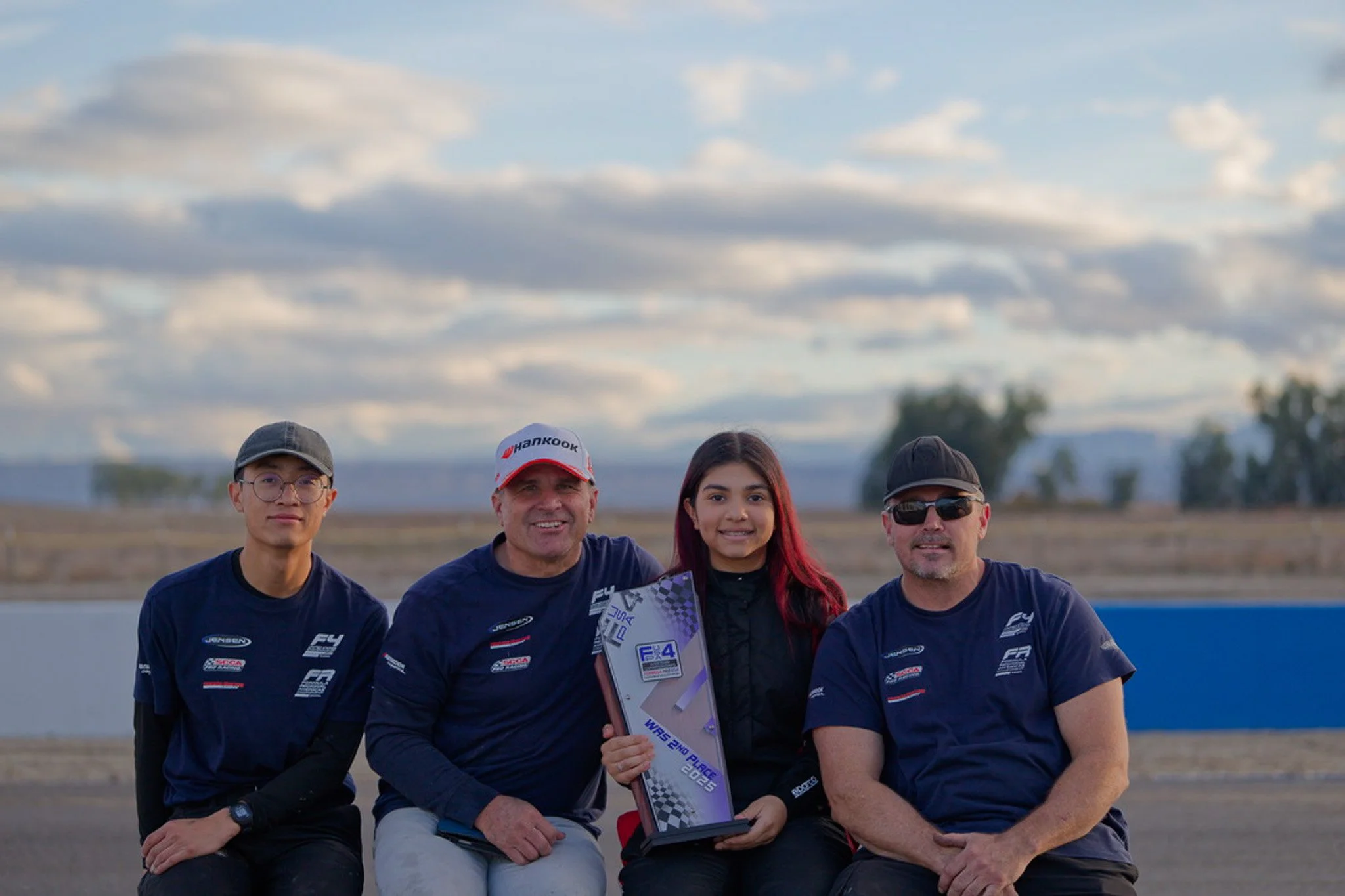 A group of five people outdoors, smiling, dressed in racing team shirts, with a cloudy sky and open field in the background. One person in the center is holding a trophy.