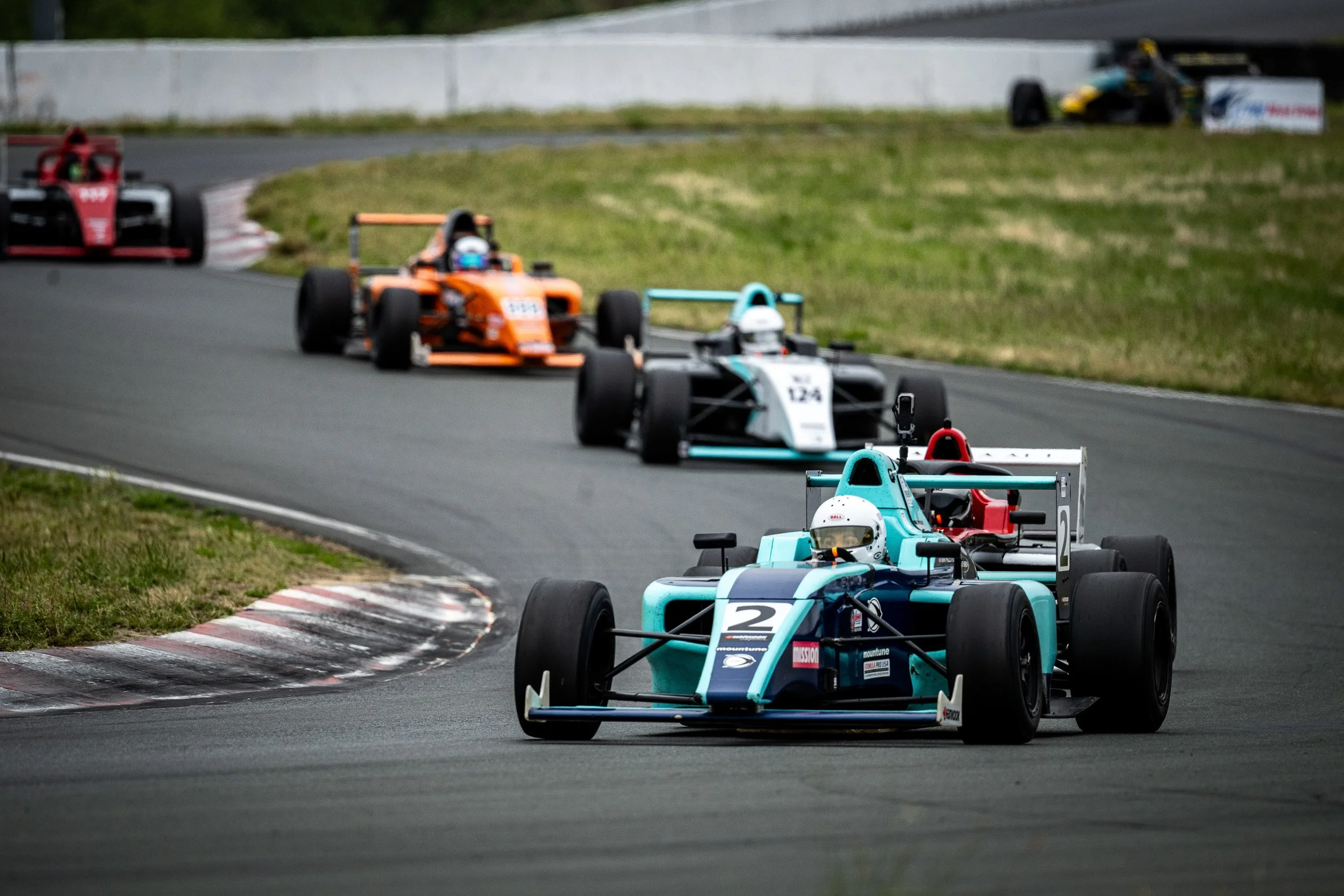 Several open-wheel race cars navigating a curve on a race track, with grass and a wall in the background.