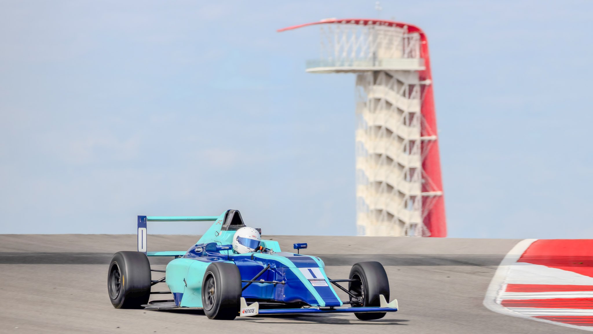 A blue race car with a driver in a helmet racing on a track, with a distinctive tower in the background.