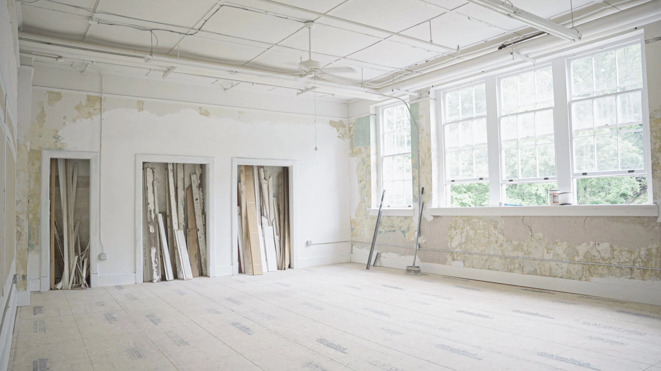 Empty room under renovation with large windows, peeling paint on walls, exposed ceiling pipes, and construction materials leaning against the wall.