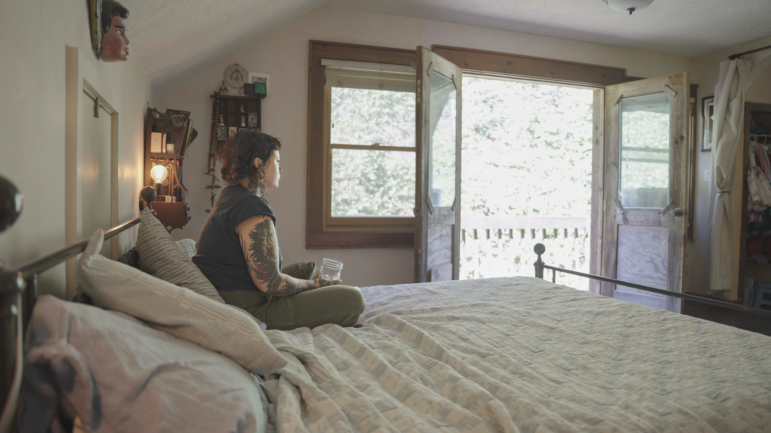 A woman with tattoos sitting on a bed in a bedroom, looking out an open door and window to a sunny outdoor balcony.