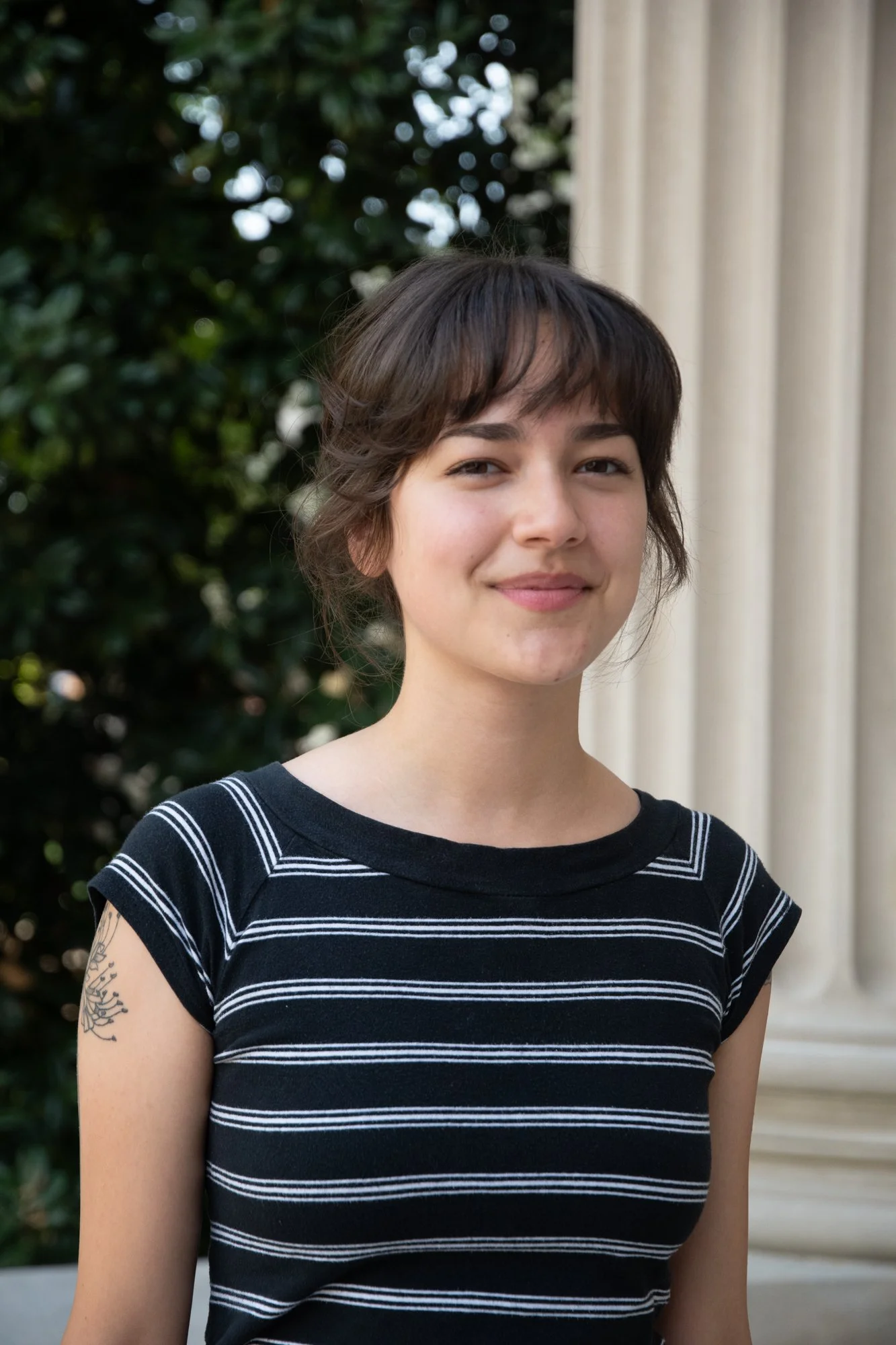 A young woman with short brown hair and light skin standing outdoors near a building with columns, wearing a black and white striped shirt, smiling softly.