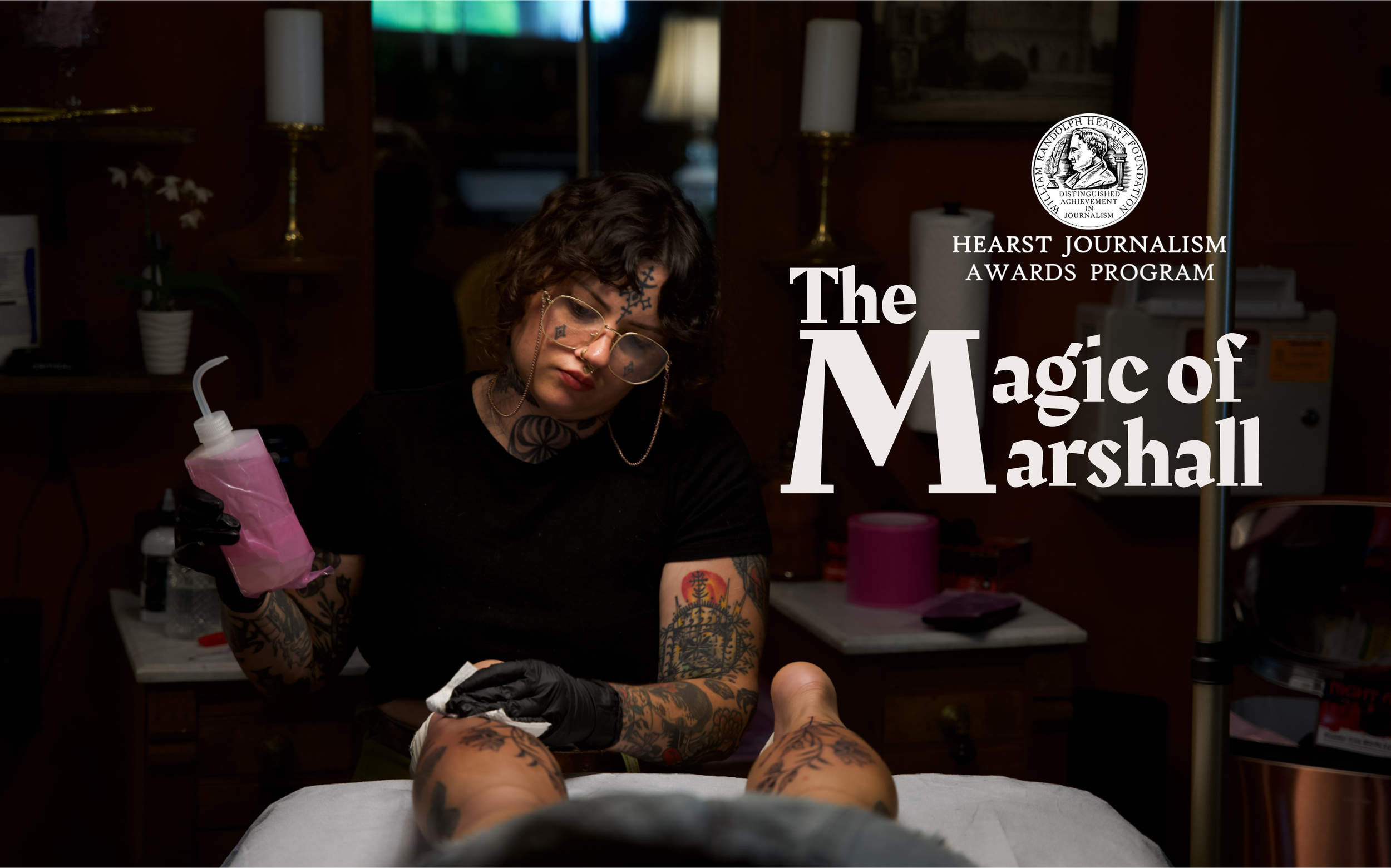Tattoo artist with glasses and tattoos working on a client's leg in a tattoo studio, with awards and art on the wall in the background.