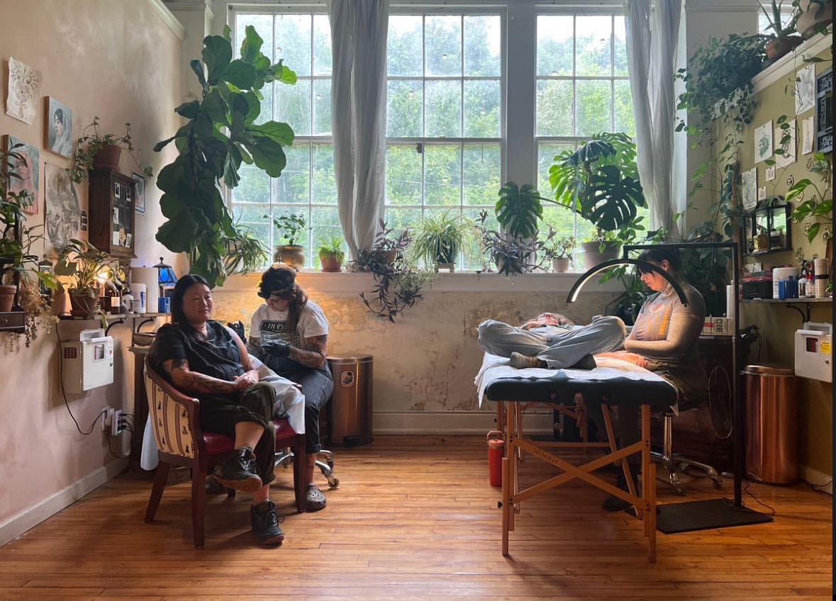 A tattoo artist sitting on a chair and working on a client with colorful tattoos inside a cozy tattoo studio filled with plants, artwork, and natural light.