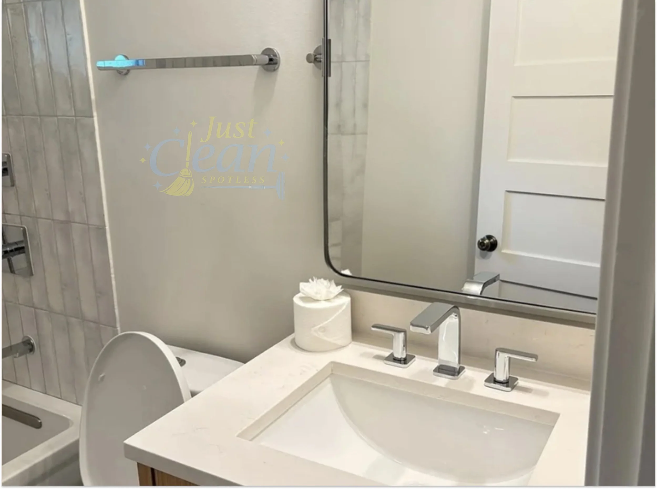 Bathroom mirror over a sink with a white countertop, a faucet, and a roll of toilet paper on a holder. A closed cabinet door and a towel bar are visible in the background. The mirror has a sticker with the words "Just Clean Spotless" and a broom illu