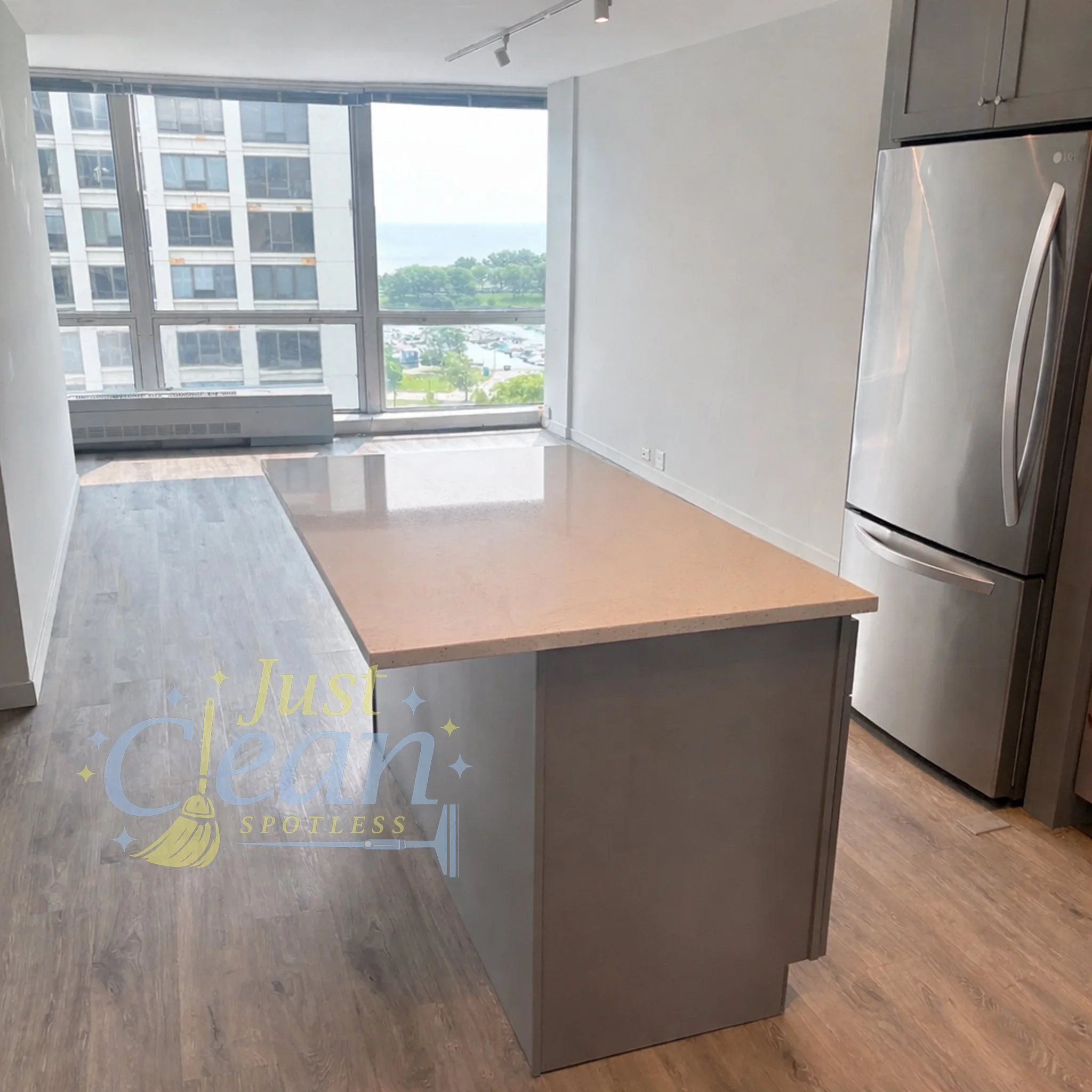 A modern apartment kitchen with large windows overlooking a city and green park, featuring a light-colored countertop and a stainless steel refrigerator.