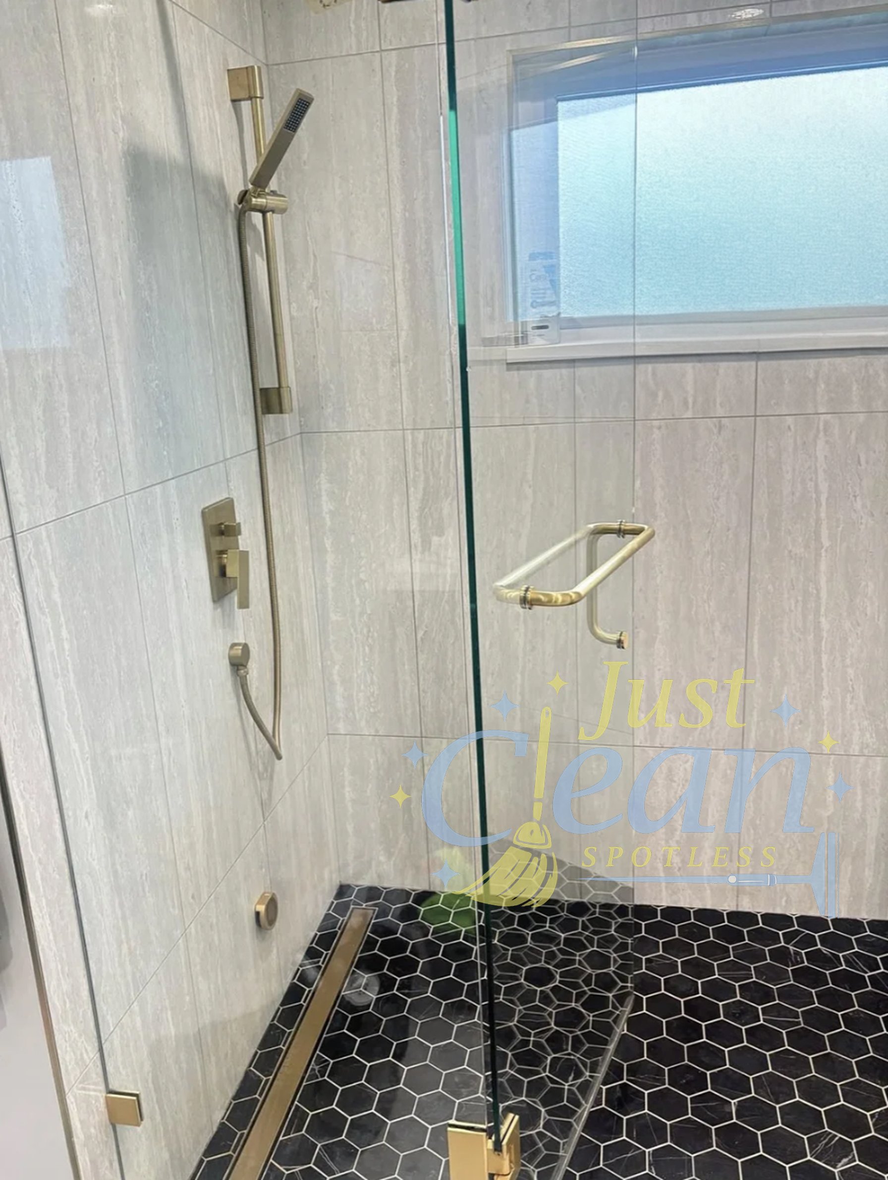 A bathroom shower with light beige tiled walls, a small window with frosted glass, a brass grab bar, a brass shower handle, a brass handrail, a soap dish, and a glass shower door with a brass handle. The shower floor is covered with black and white h