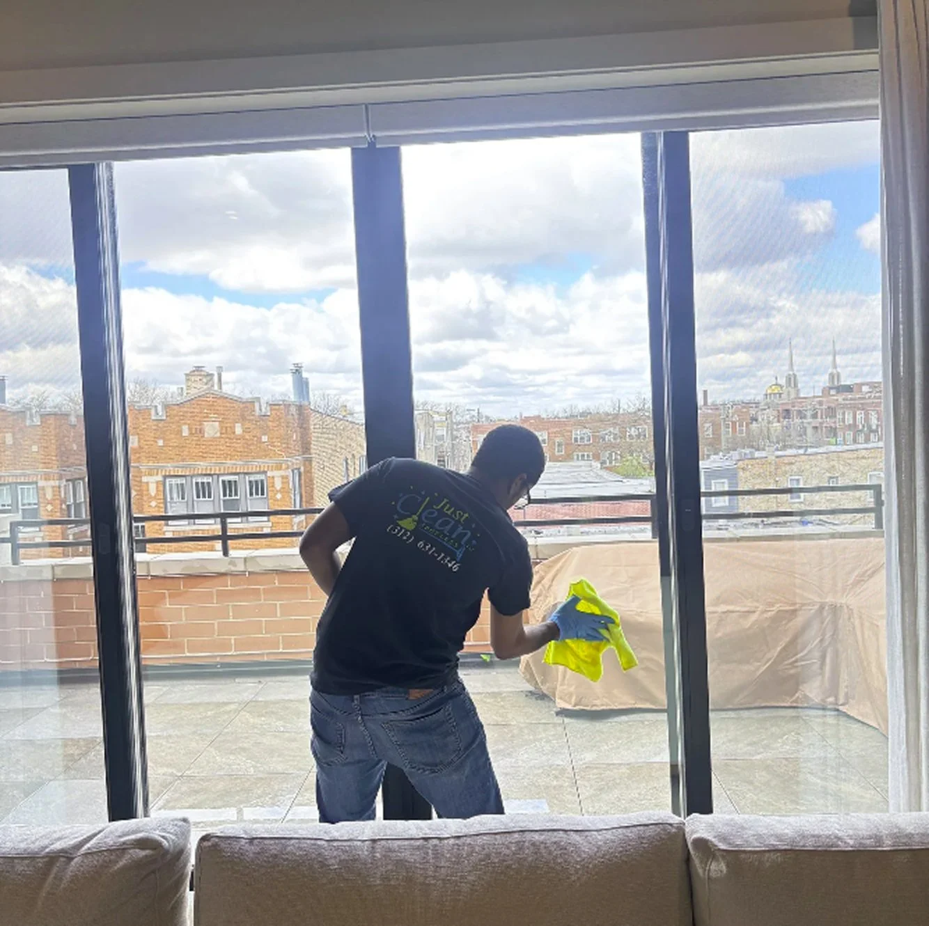 A person cleaning a glass sliding door on a balcony, with city buildings and a cloudy sky in the background.