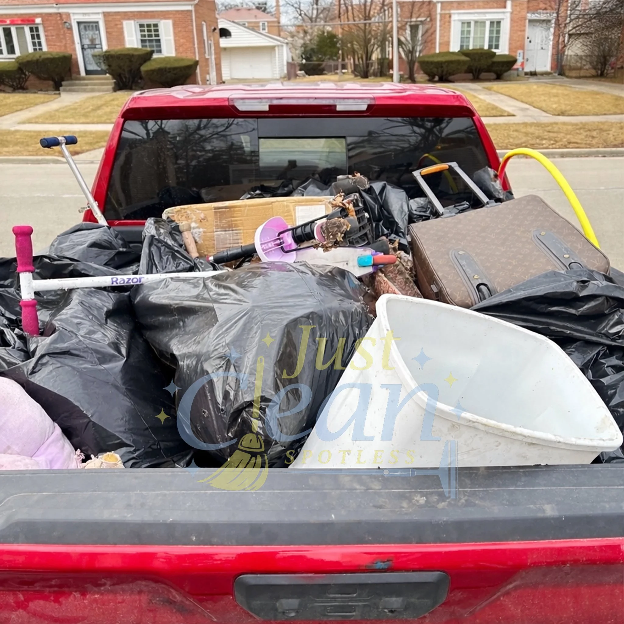 The image shows the back of a red vehicle with various items piled in the truck bed, including black trash bags, a pink shovel, and other miscellaneous objects. The background features residential houses with brick exteriors, windows with white shutters, steps leading to the doors, and well-maintained front yards.