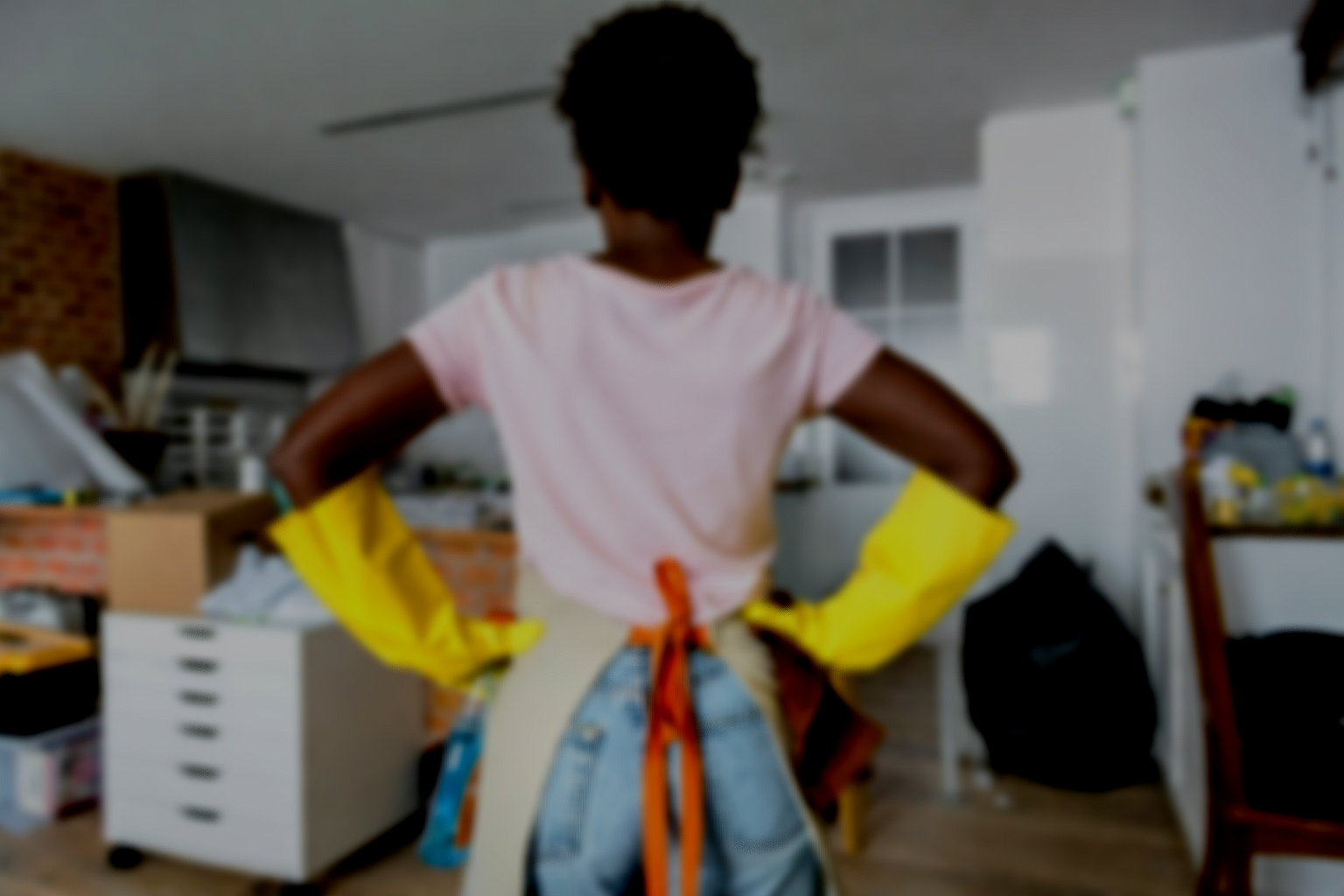 Person standing in a kitchen with hands on hips, wearing yellow rubber gloves, pink shirt, and apron, facing away.