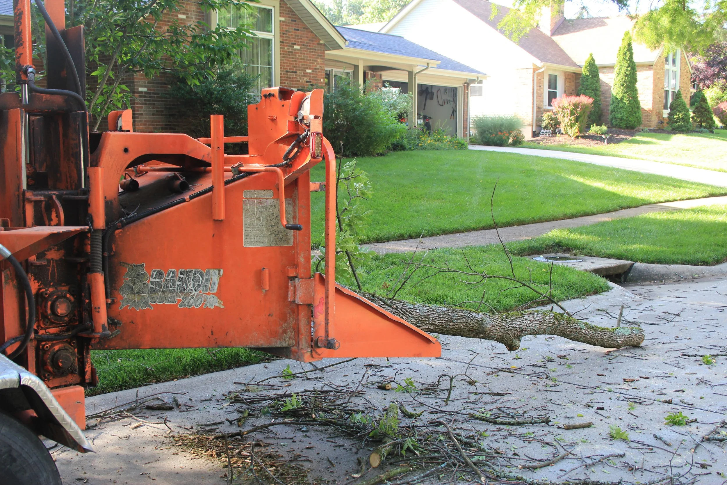 A large orange woodchipper machine on a driveway with tree branch debris from tree removal clean up and a sidewalk in a residential neighborhood with houses, trees, and a lawn.