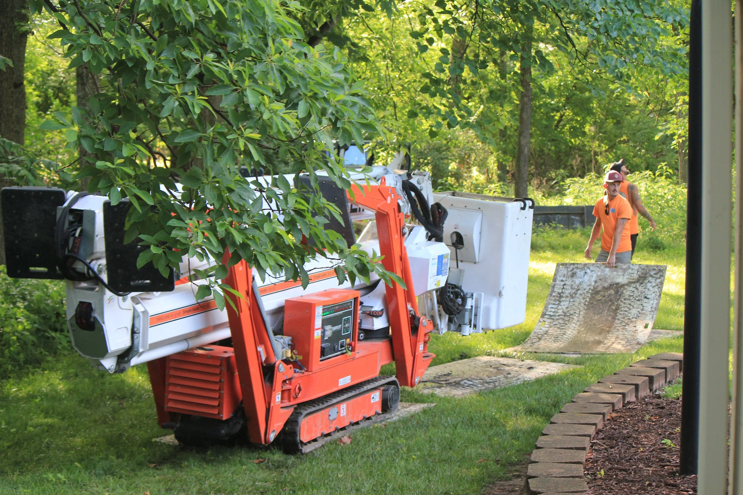 A construction site with a focus on a large orange and white stormwater treatment device, surrounded by green trees. Two workers wearing orange shirts and hats are visible in the background, one standing and the other walking behind.