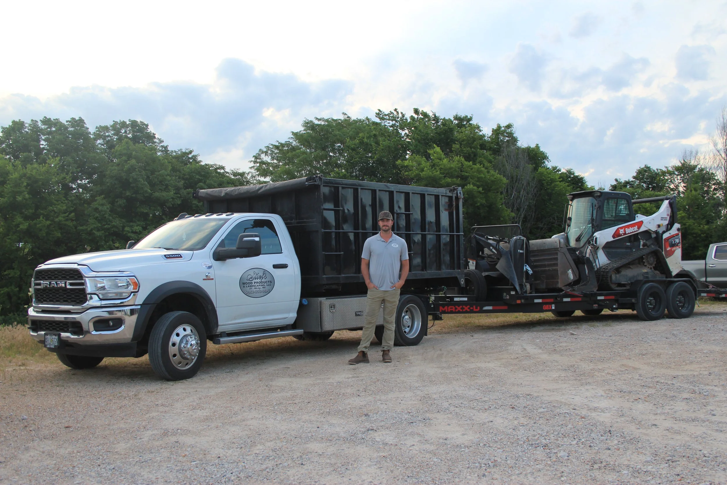 A man standing in front of a white pickup truck with a black flatbed trailer attached, carrying construction equipment, in an outdoor area with trees and a cloudy sky.