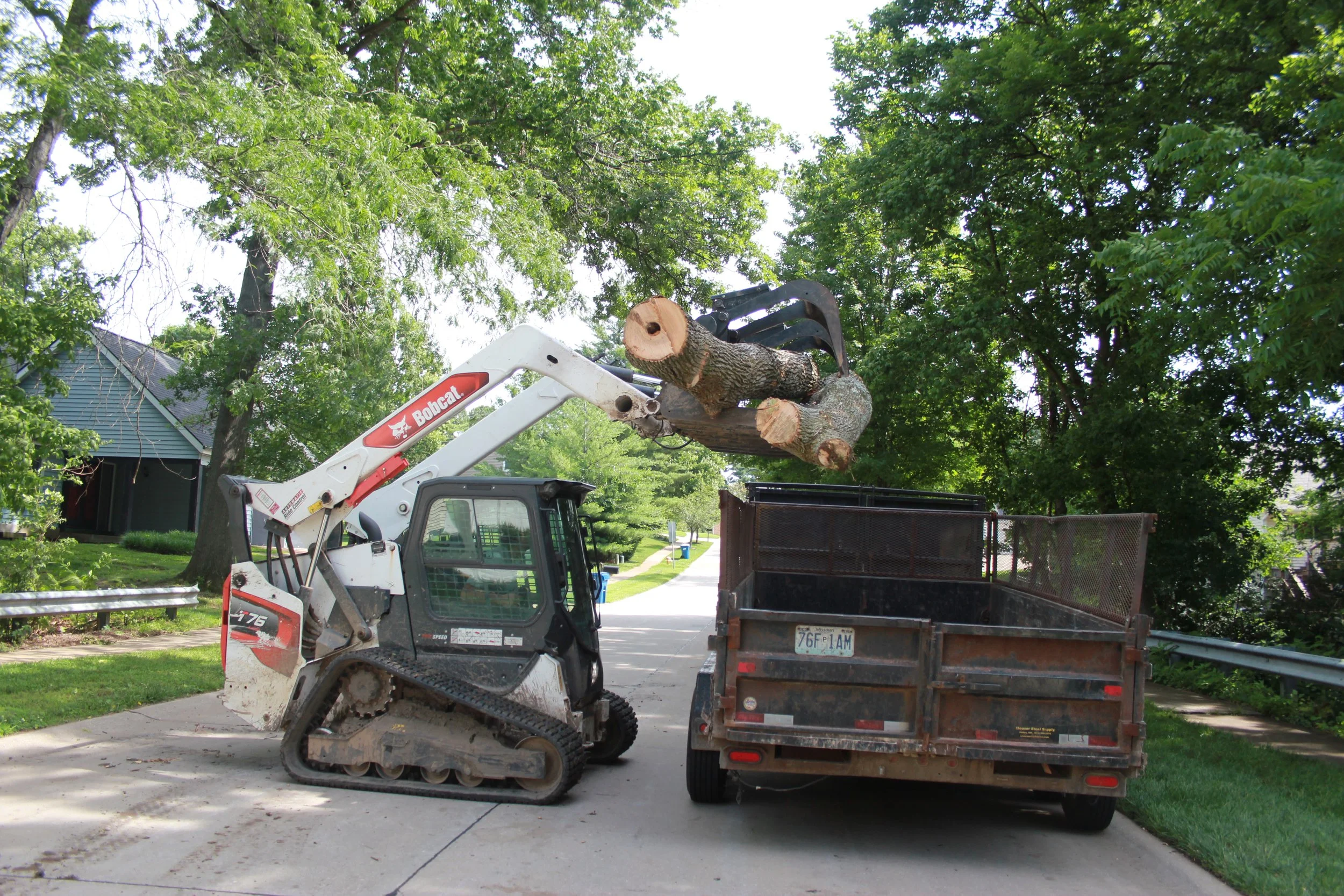 A Bobcat skid steer loader lifting a large cut log into a black dump truck parked on a residential street with green trees and houses in the background.
