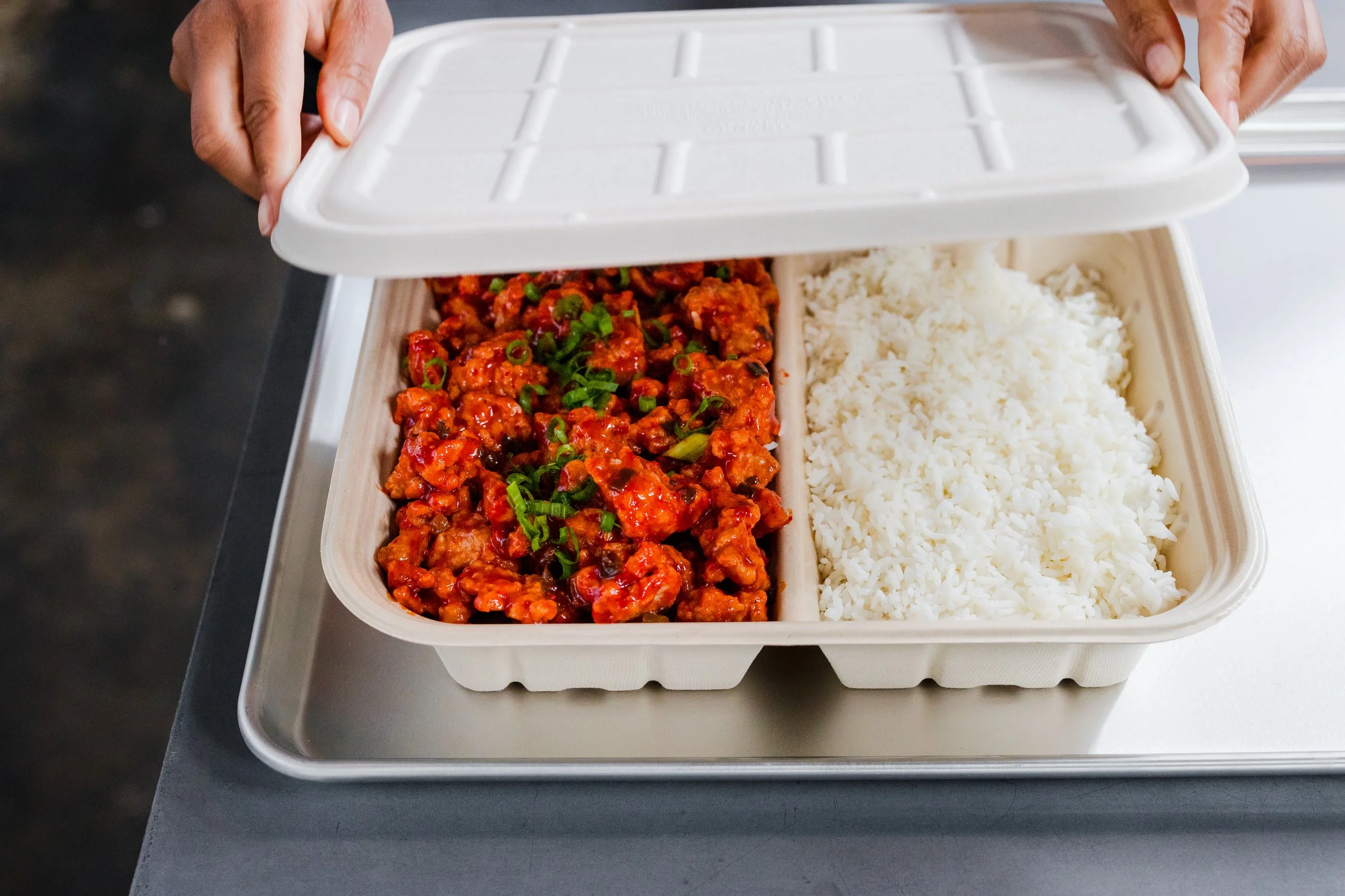 Container of Chinese takeout with orange chicken garnished with green onions and white rice inside.