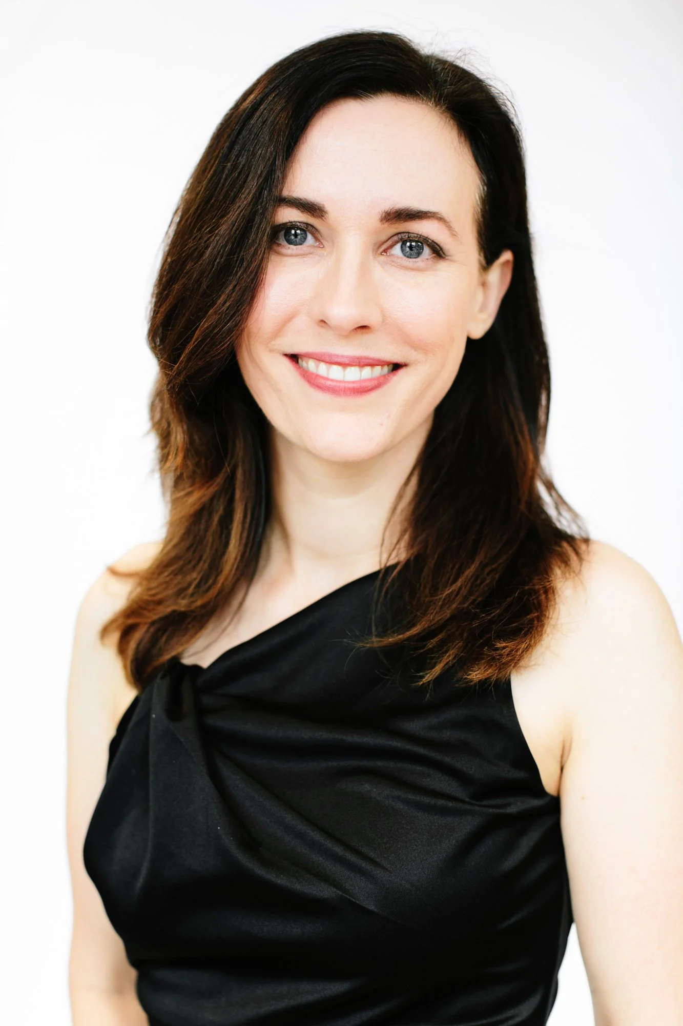 A woman with dark brown hair, blue eyes, and fair skin, smiling, wearing a black sleeveless dress with a knot detail on the shoulder, against a white background.