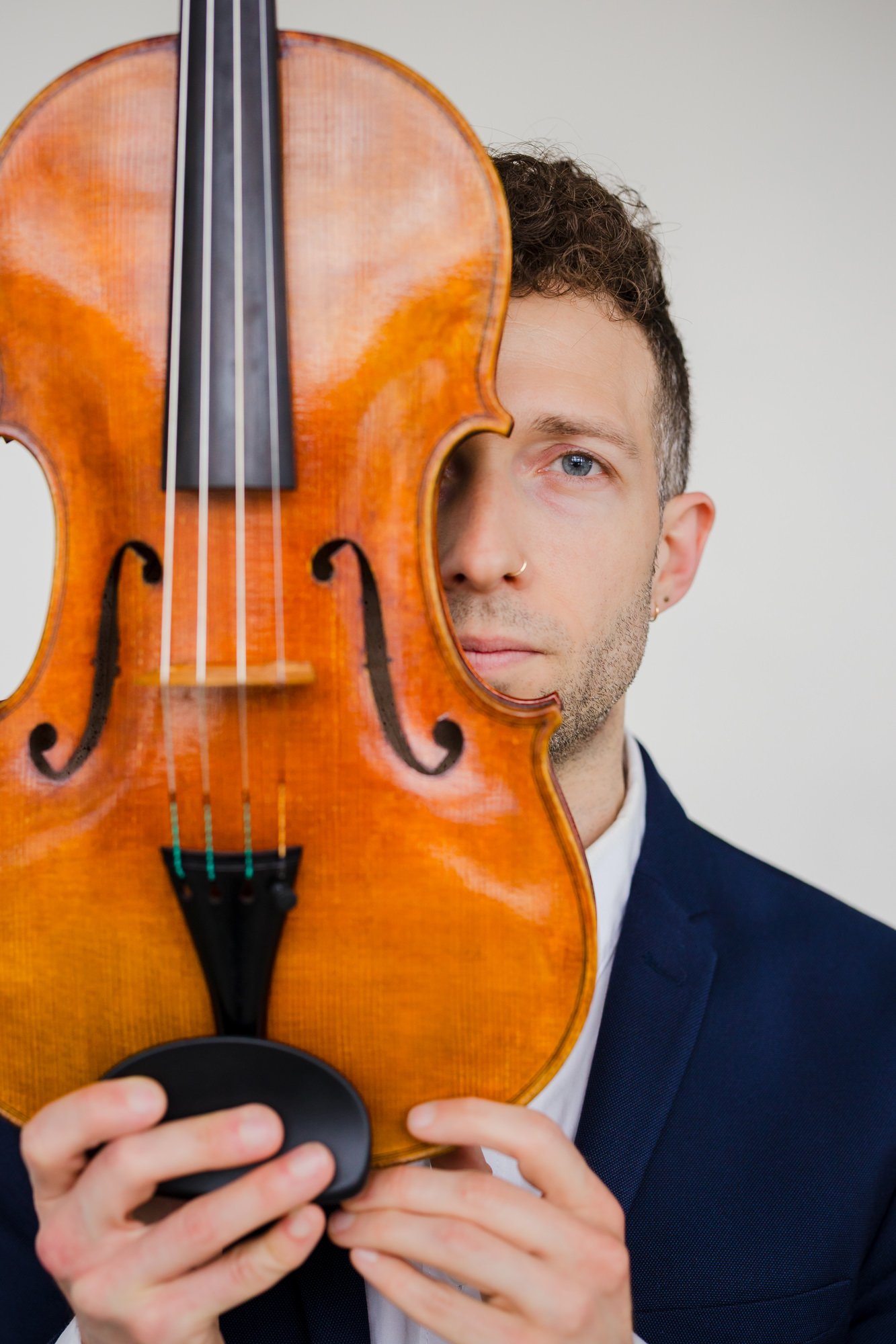 A man with blue eyes, short curly hair, wearing a navy blazer and white shirt, holding a violin in front of his face, partially obscuring his face, with the background being a plain white wall.