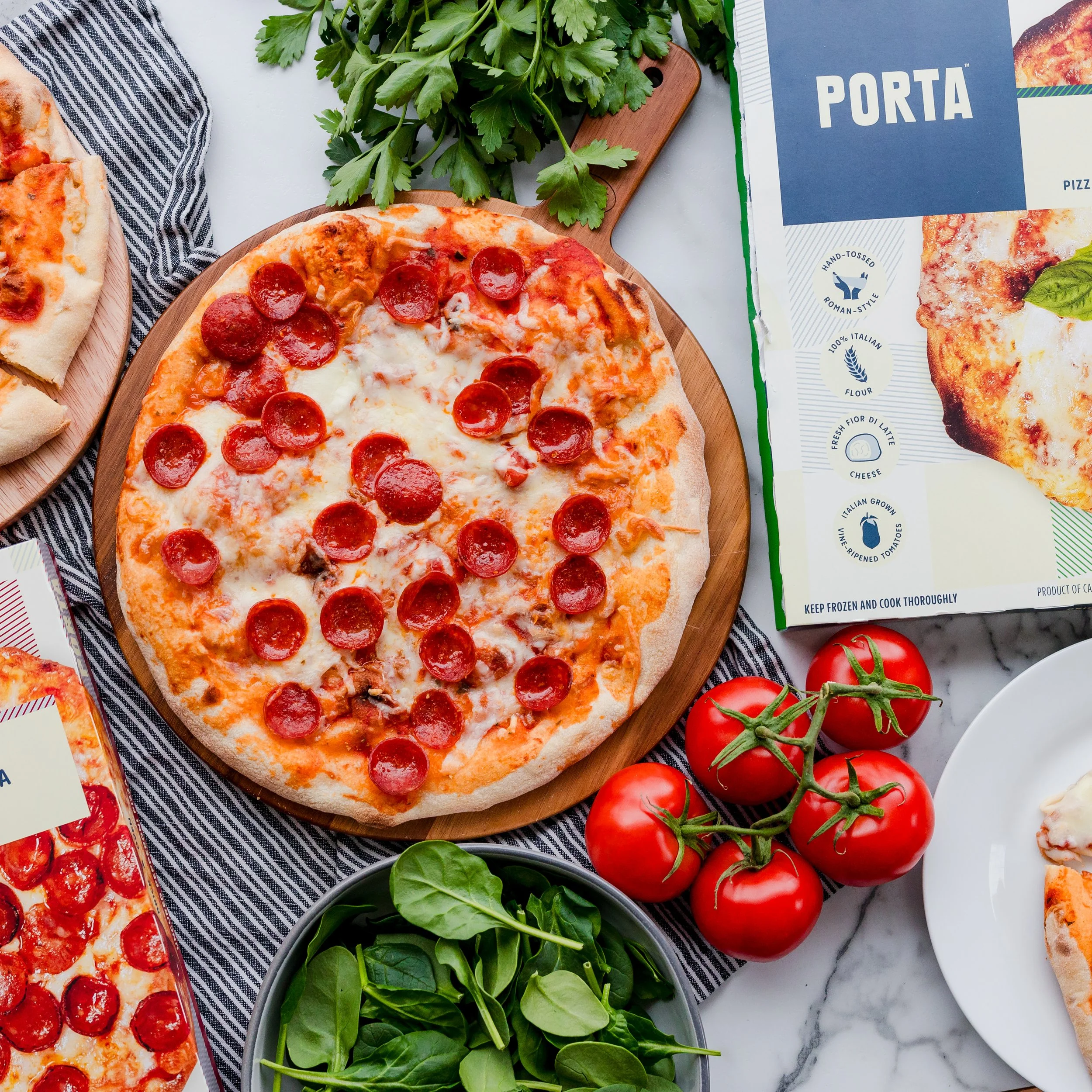 A pepperoni pizza on a round wooden pizza peel, surrounded by fresh tomatoes, basil, and pizza boxes.