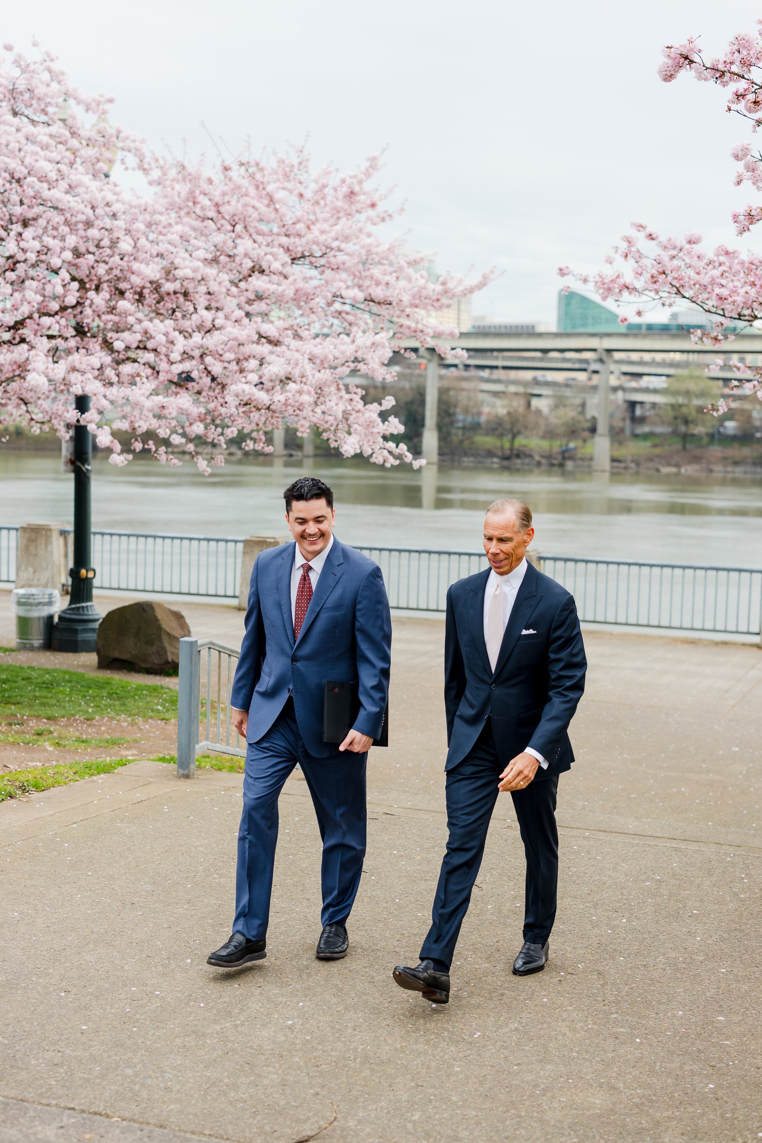 Two men in suits walking and talking outdoors near a waterfront with cherry blossom trees in bloom.