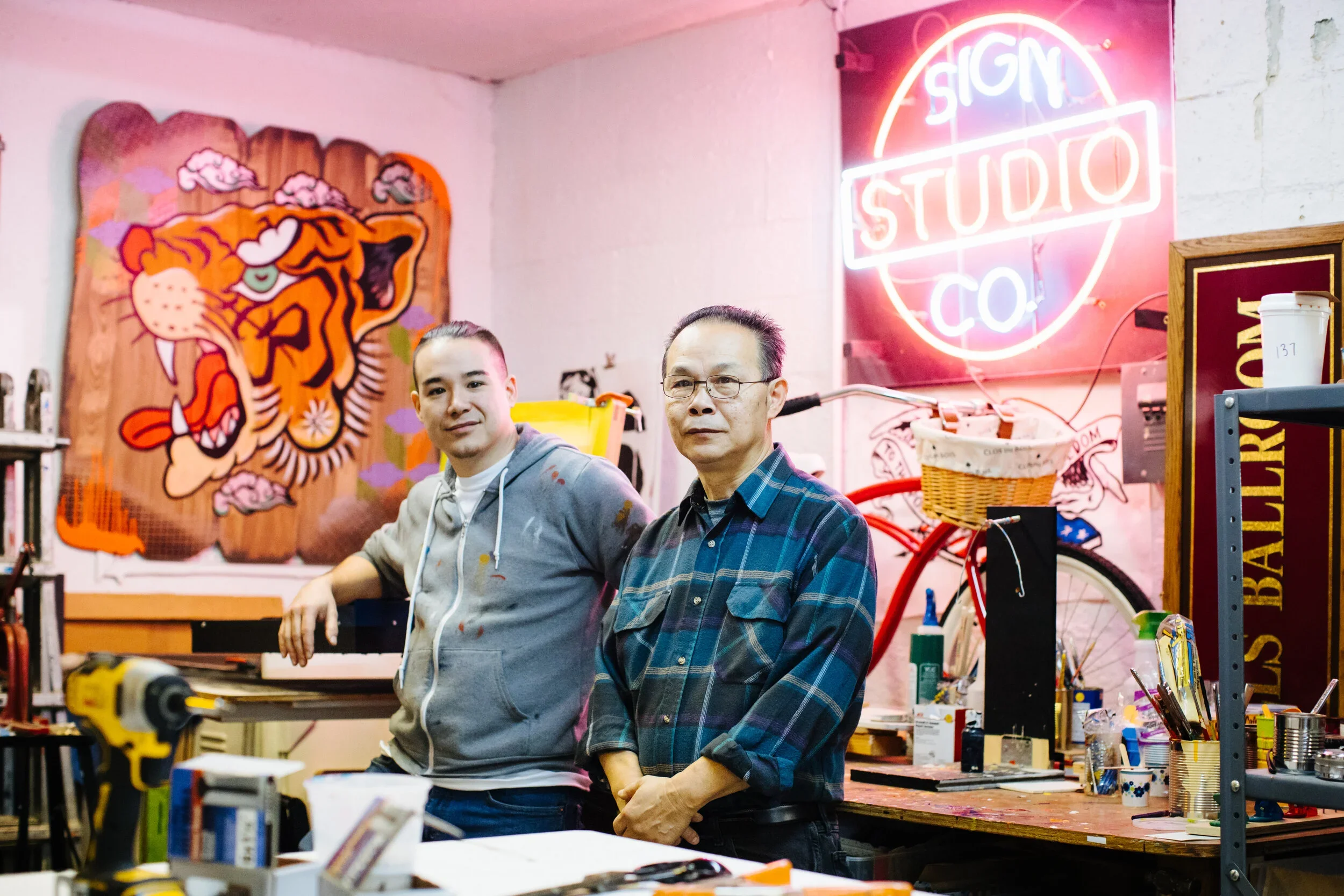 Two men in a workshop with a neon sign that says 'SIGN STUDIO CO', a tiger portrait on the wall, and various tools and crafts on the workbenches.