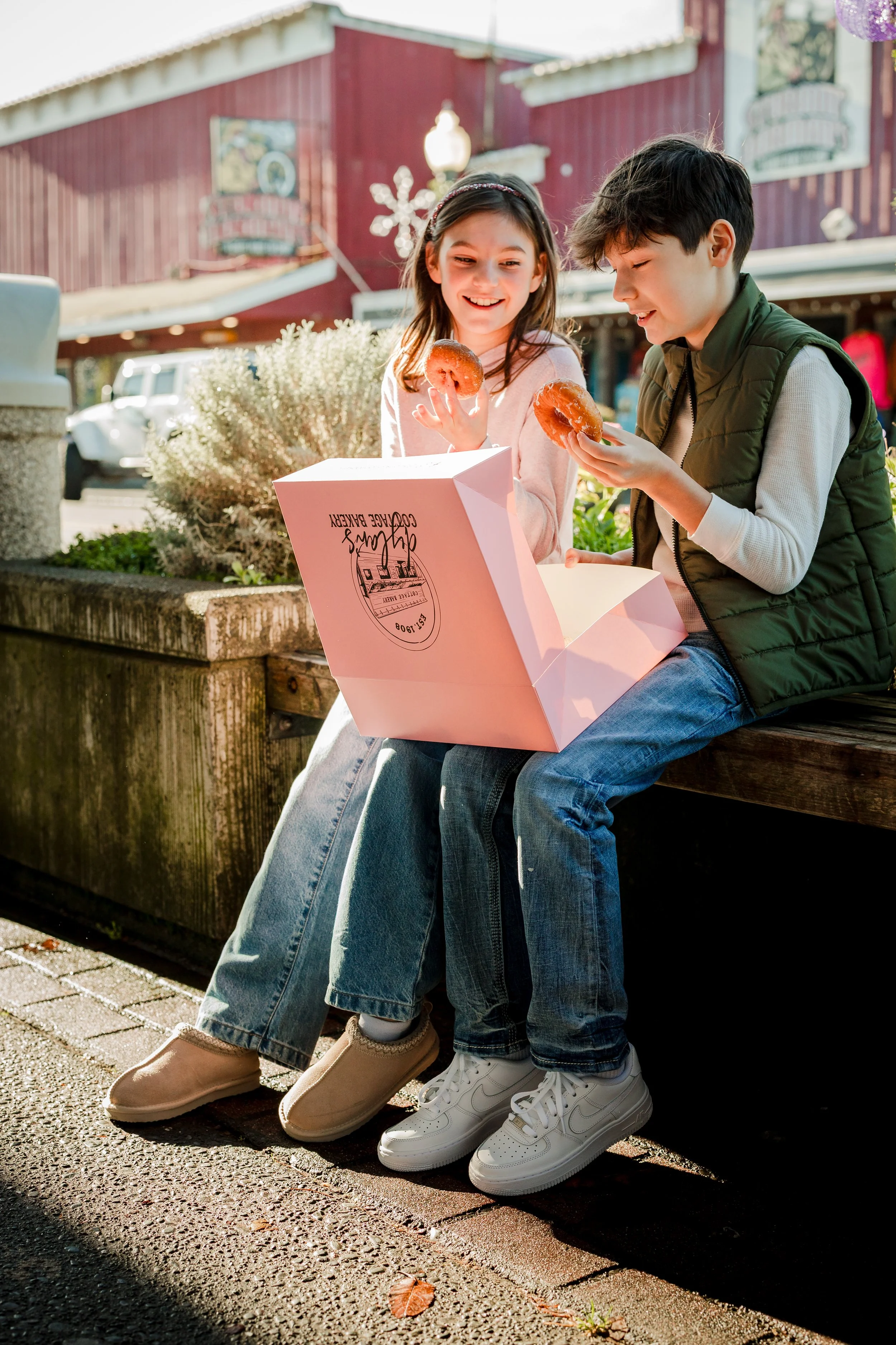 A boy and girl sitting on a park bench outdoors, sharing a pink bakery box and eating donuts, with a red building and parked cars in the background.