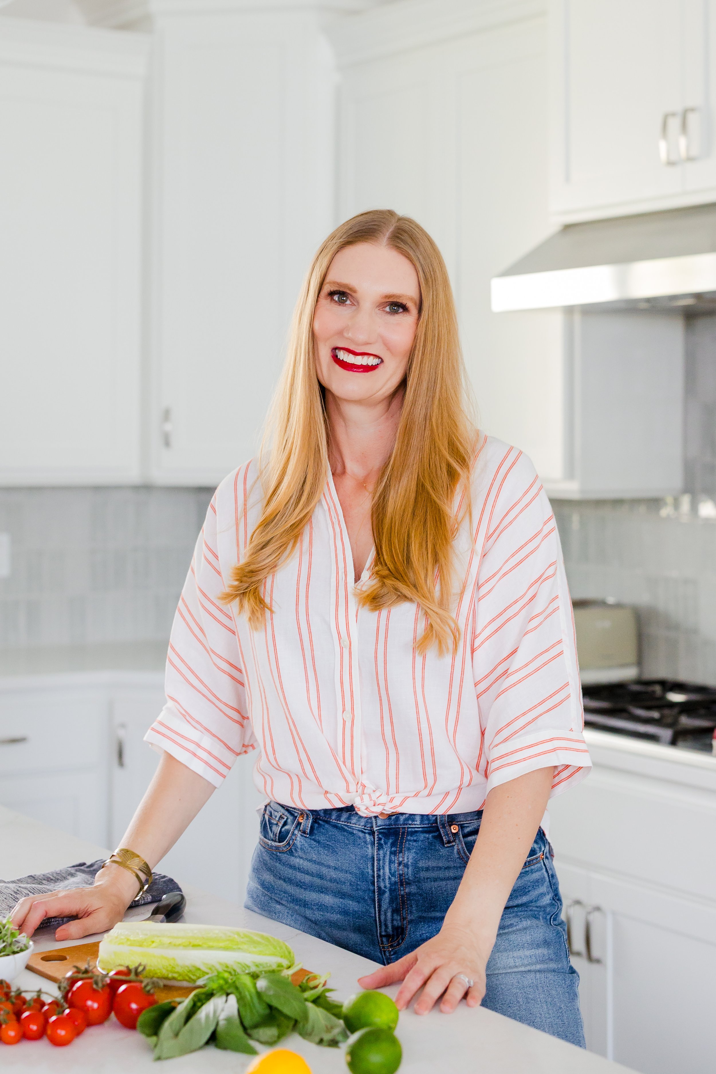 A woman with long red hair, wearing a white shirt with red stripes, stands in a bright kitchen with white cabinets. She is smiling and leaning on a kitchen counter that has various fresh vegetables including tomatoes, lettuce, and limes.
