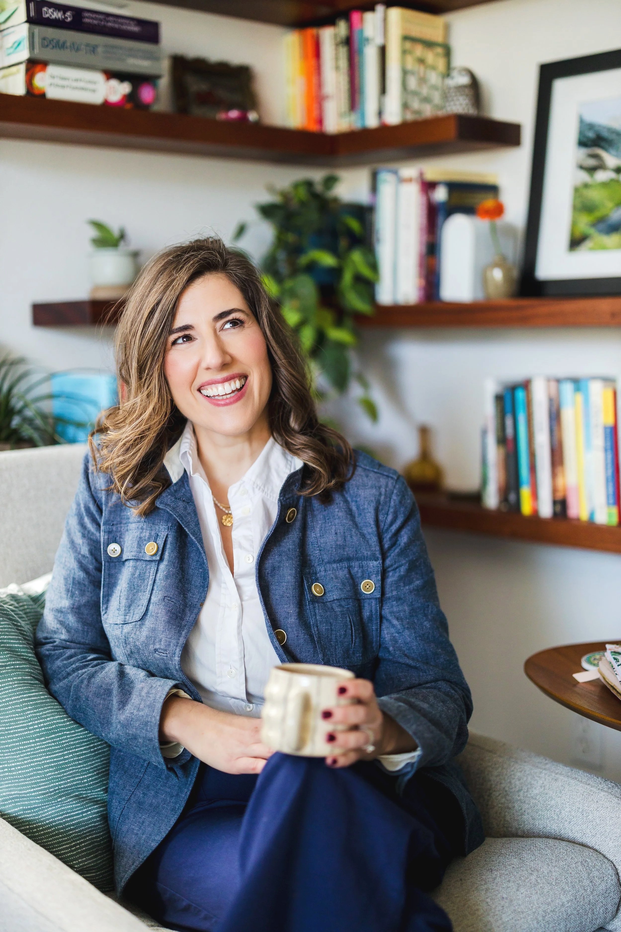 A woman with shoulder-length brown hair, wearing a denim jacket and white shirt, sitting on a beige couch in a living room, smiling while holding a mug. Behind her are bookshelves with colorful books and a framed picture, and some houseplants.