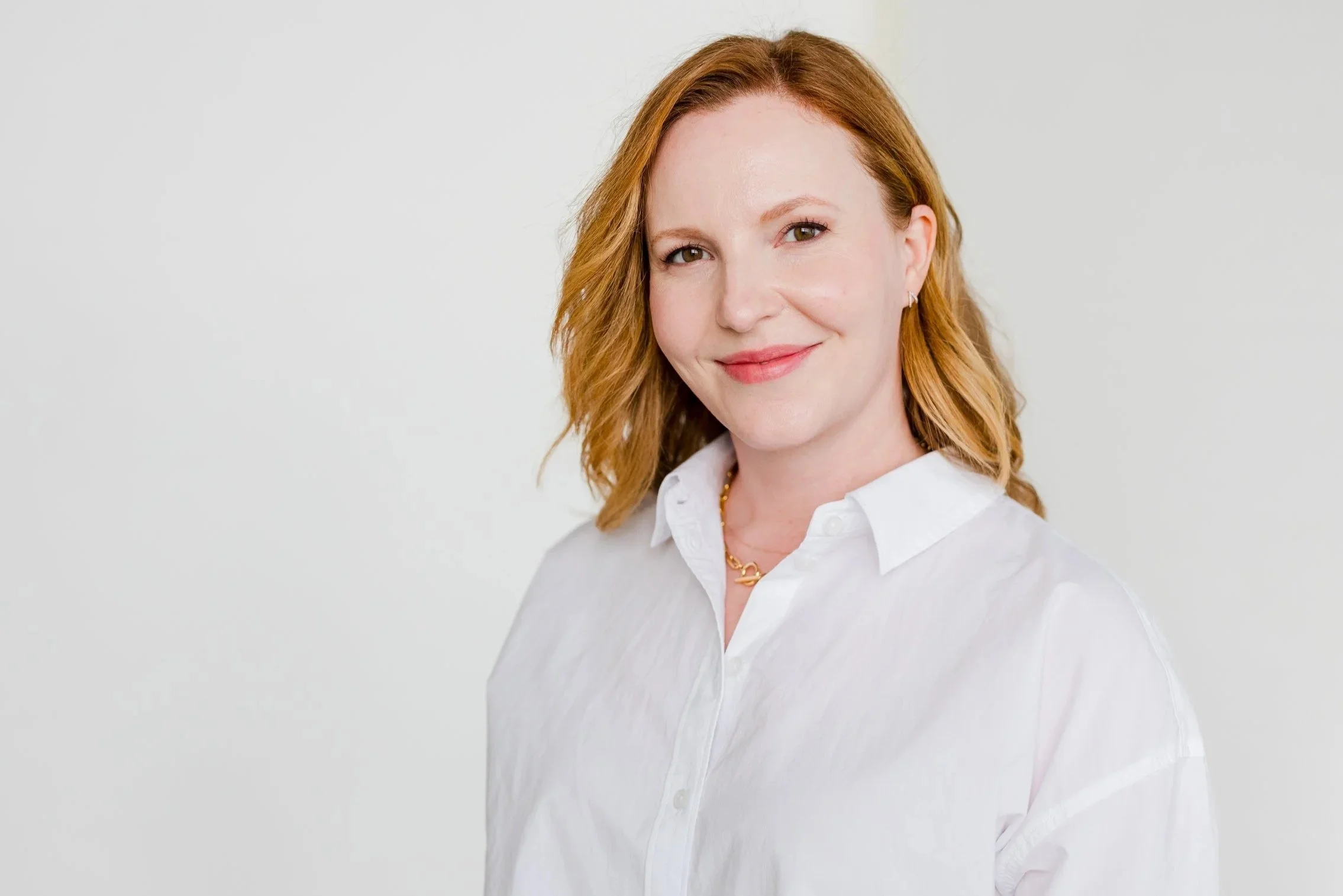 A woman with red hair, wearing a white button-up shirt, smiling softly, standing against a plain light background.