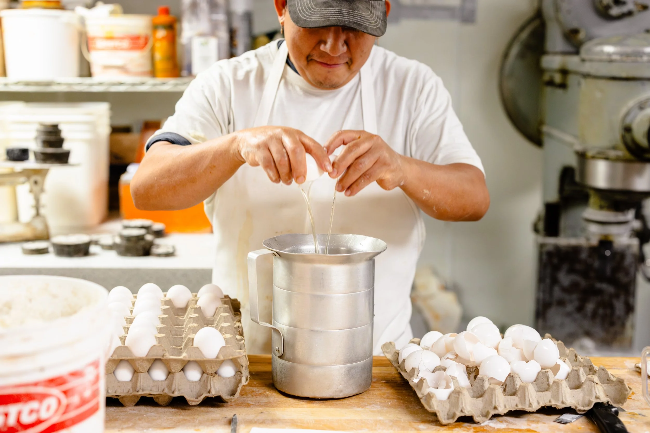 A person cracking eggs into a metal bowl in a kitchen or bakery setting, with egg cartons and various kitchen supplies visible around.