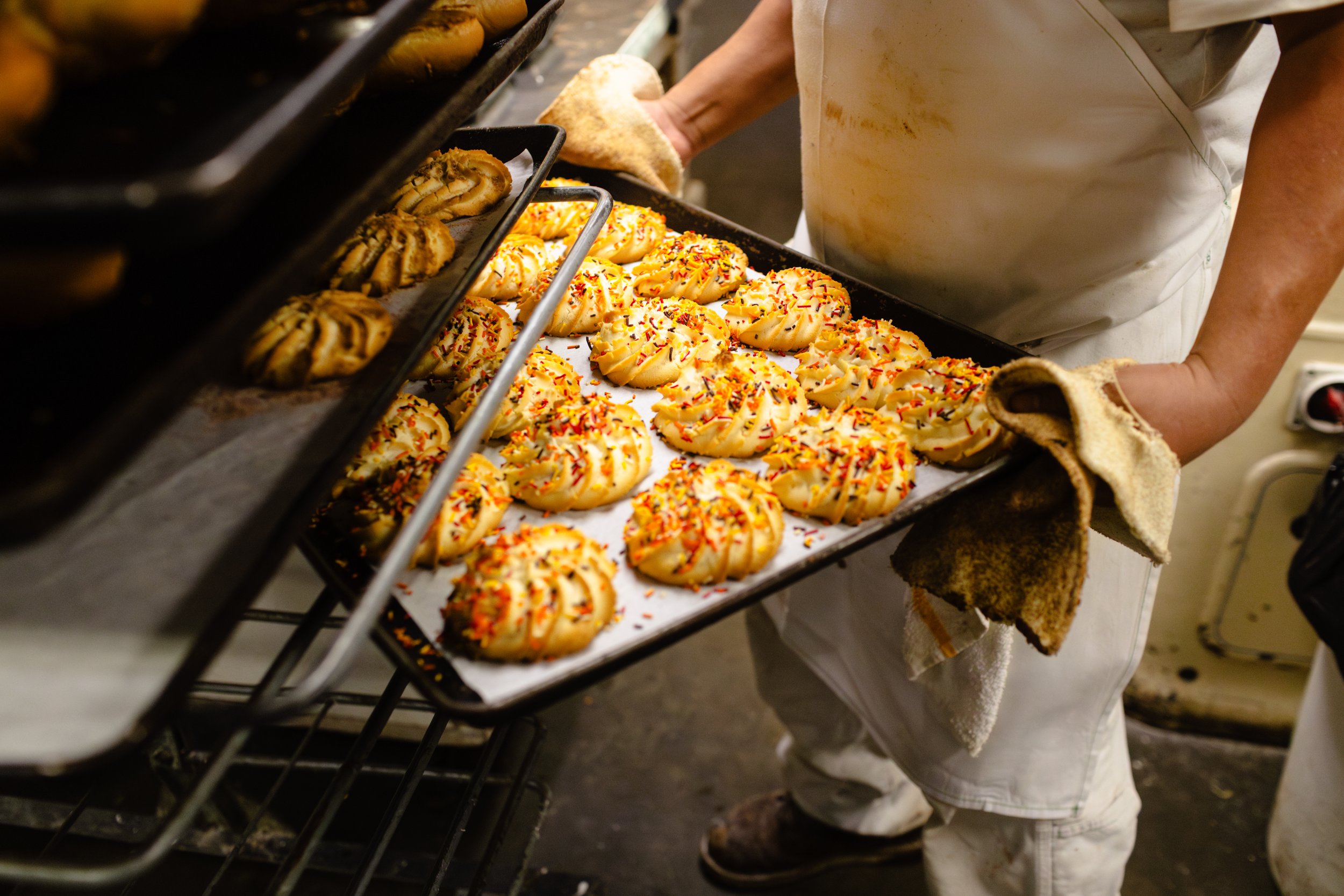Baker wearing oven mitts removing a tray of decorated cookies with rainbow sprinkles from an oven in a bakery.