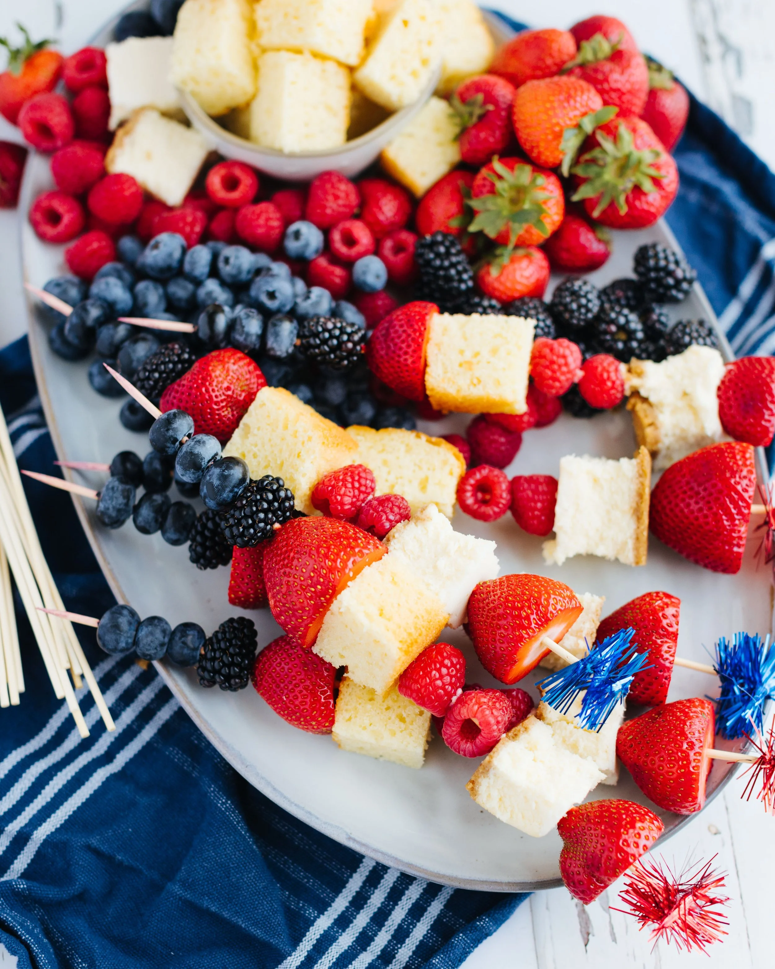A platter of fresh strawberries, blueberries, blackberries, raspberries, sponge cake pieces, and skewers with strawberries and blueberries.