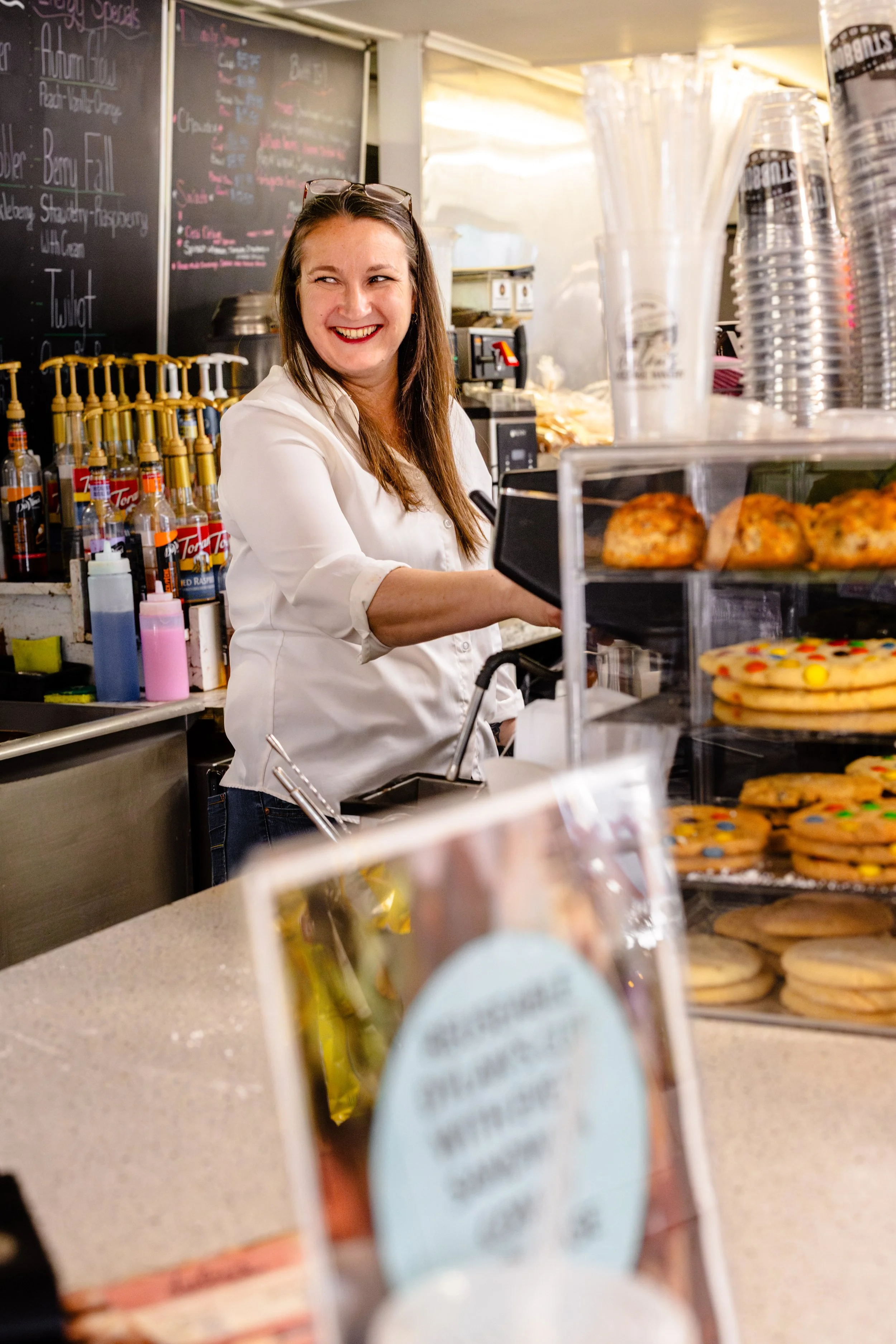 A woman with glasses on her head, wearing a white shirt, is working behind the counter of a bakery or coffee shop, smiling. There are baked goods like cookies and pastries on display in front of her, and bottles of syrup or toppings are visible on th