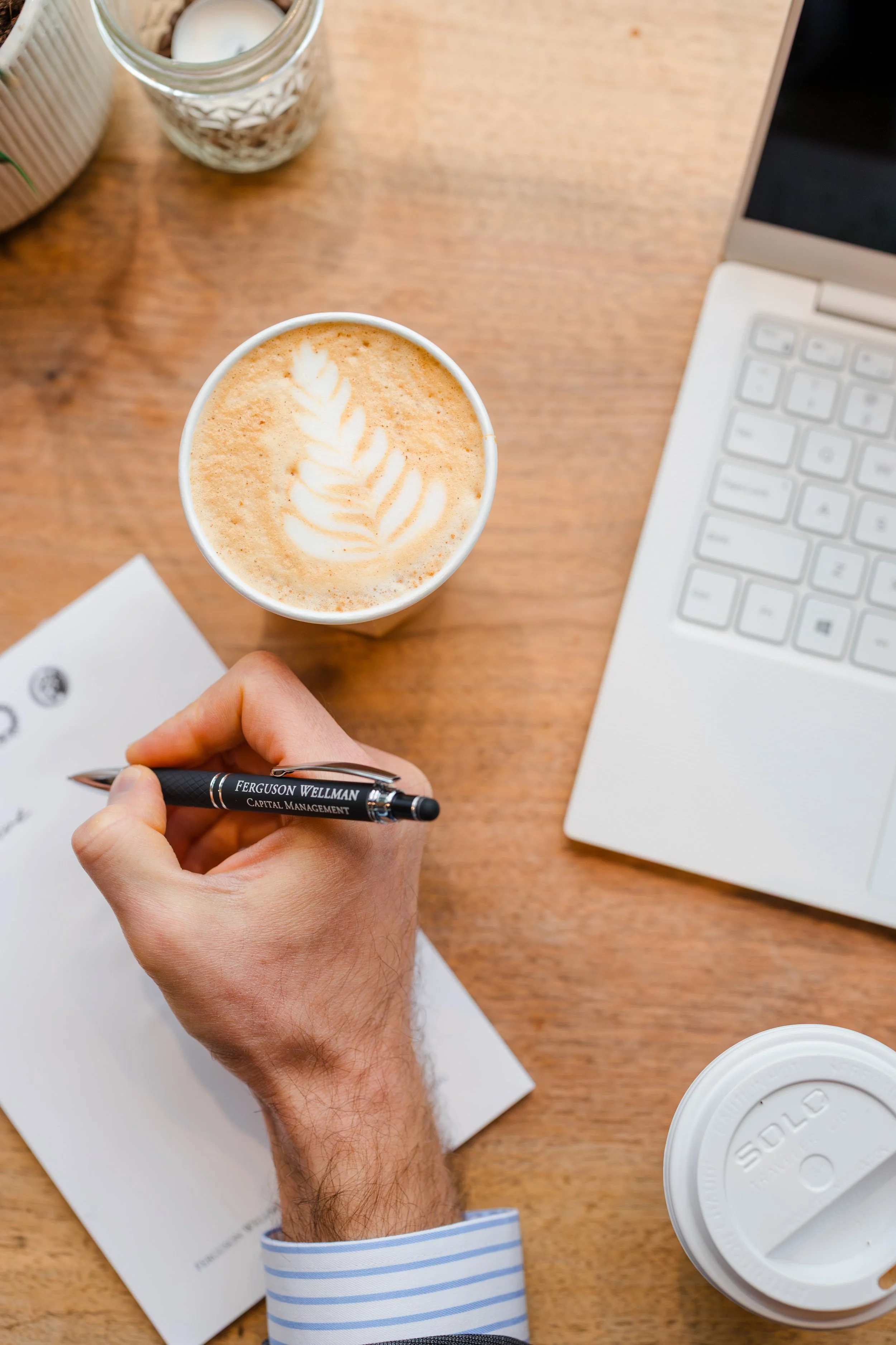 Desk with a coffee cup with latte art, a person's hand holding a pen, a blank sheet of paper, a laptop, a glass jar with a candle, and a to-go coffee cup.