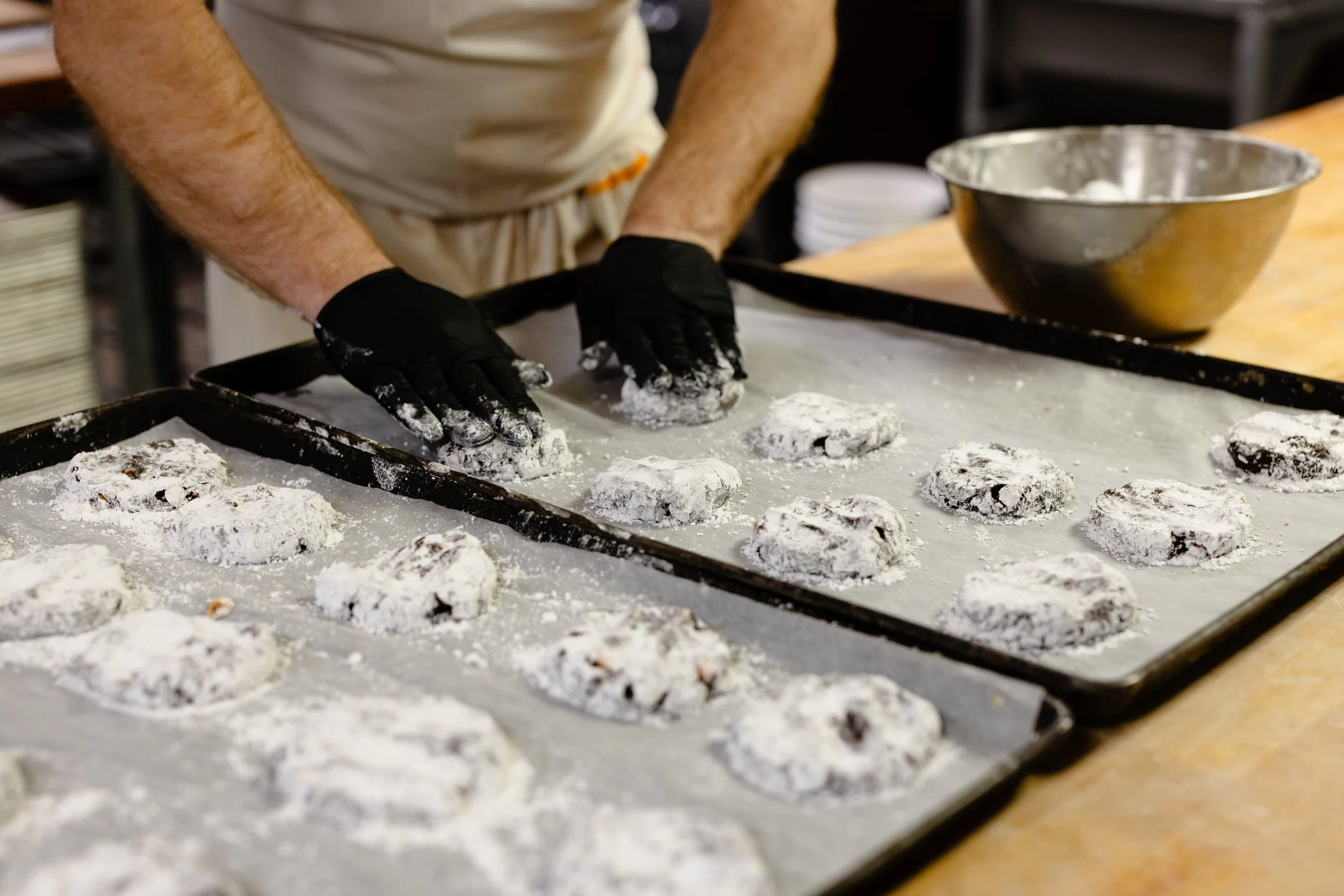 A baker wearing black gloves and a beige apron shaping chocolate chip cookies on a baking sheet dusted with flour in a bakery kitchen.