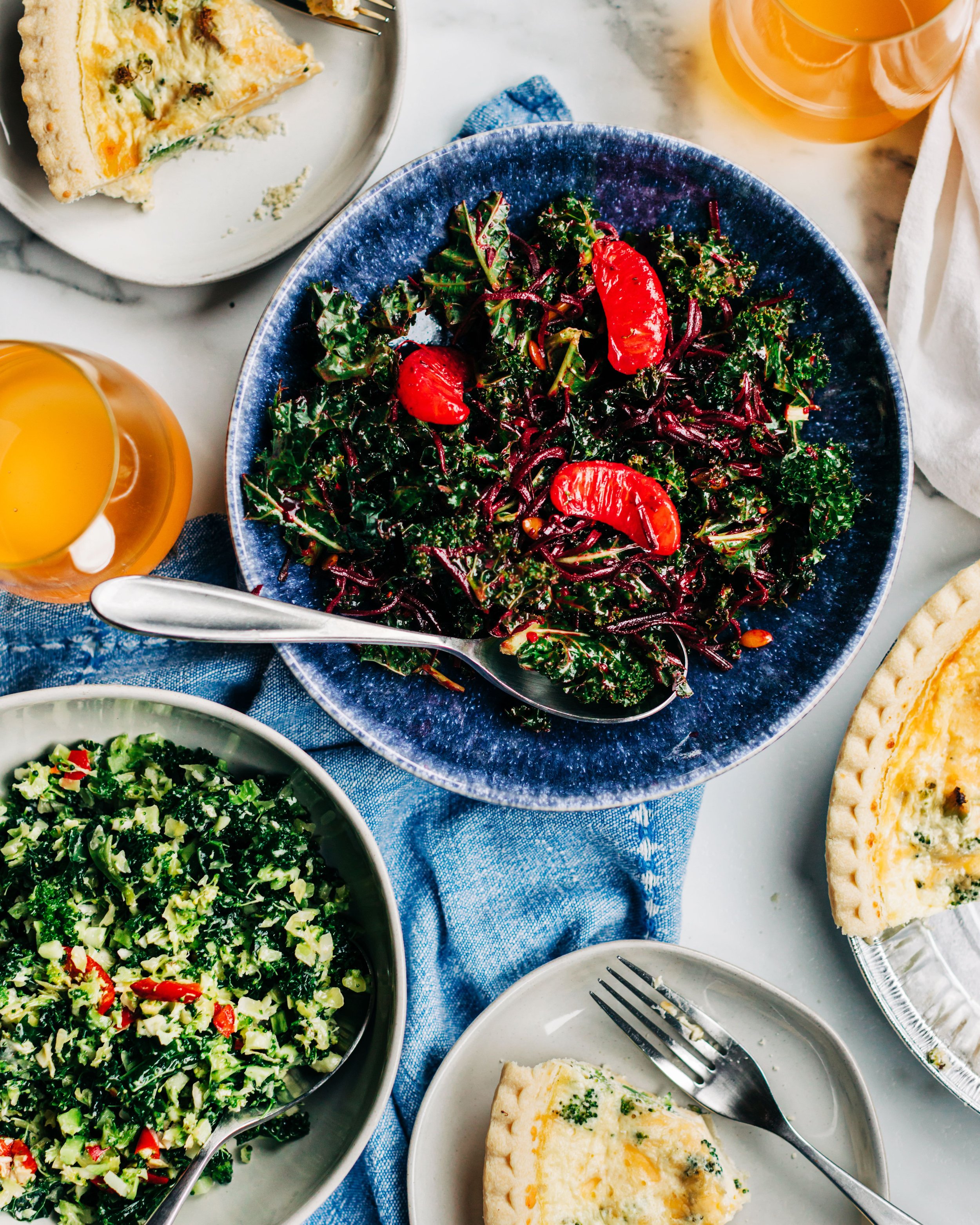 A top-down view of a dinner table with a blue plate of kale salad with cranberries, a bowl of chopped greens with red pepper, slices of quiche, and glasses of orange juice.