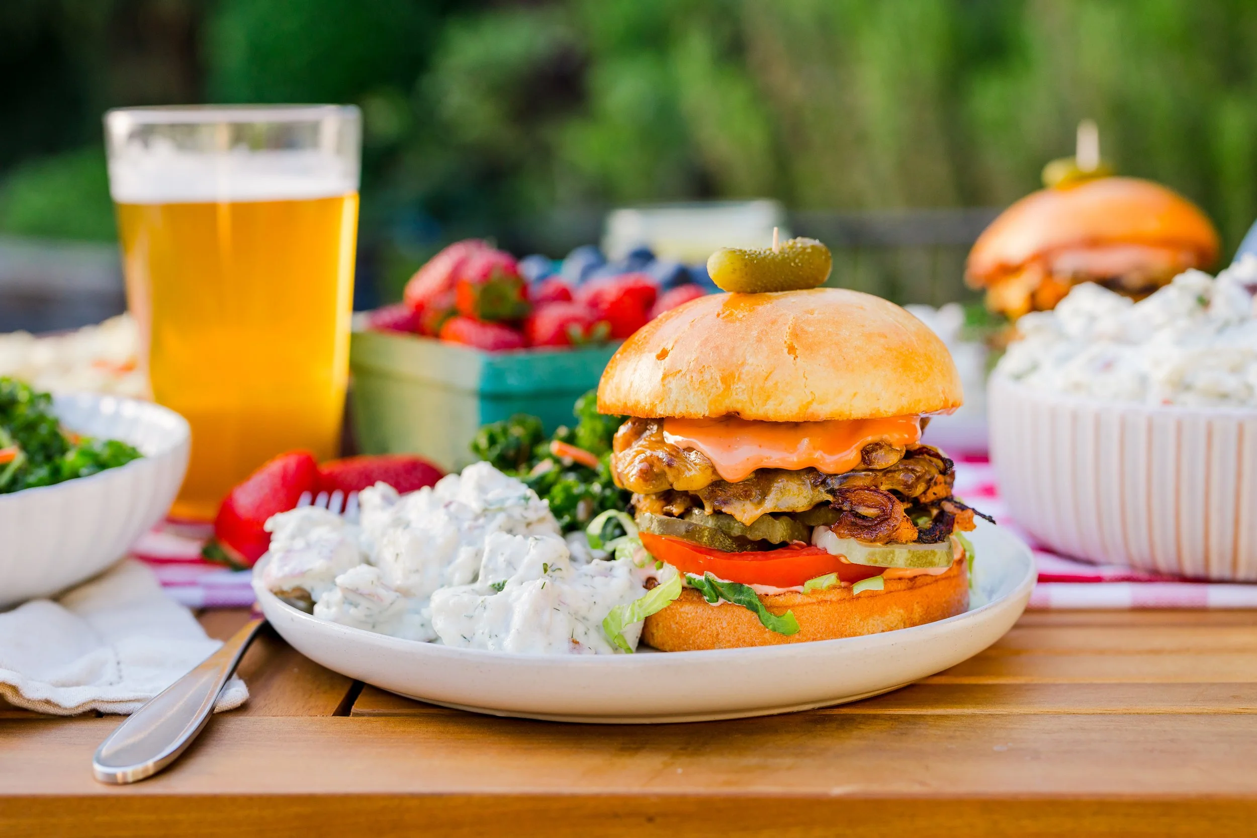 A plate with a large burger sandwich containing lettuce, tomato, pickles, bacon, cheese, and a sauce, garnished with a pickle on top, served alongside potato salad, with a glass of beer and a bowl of strawberries in the background on a wooden table o
