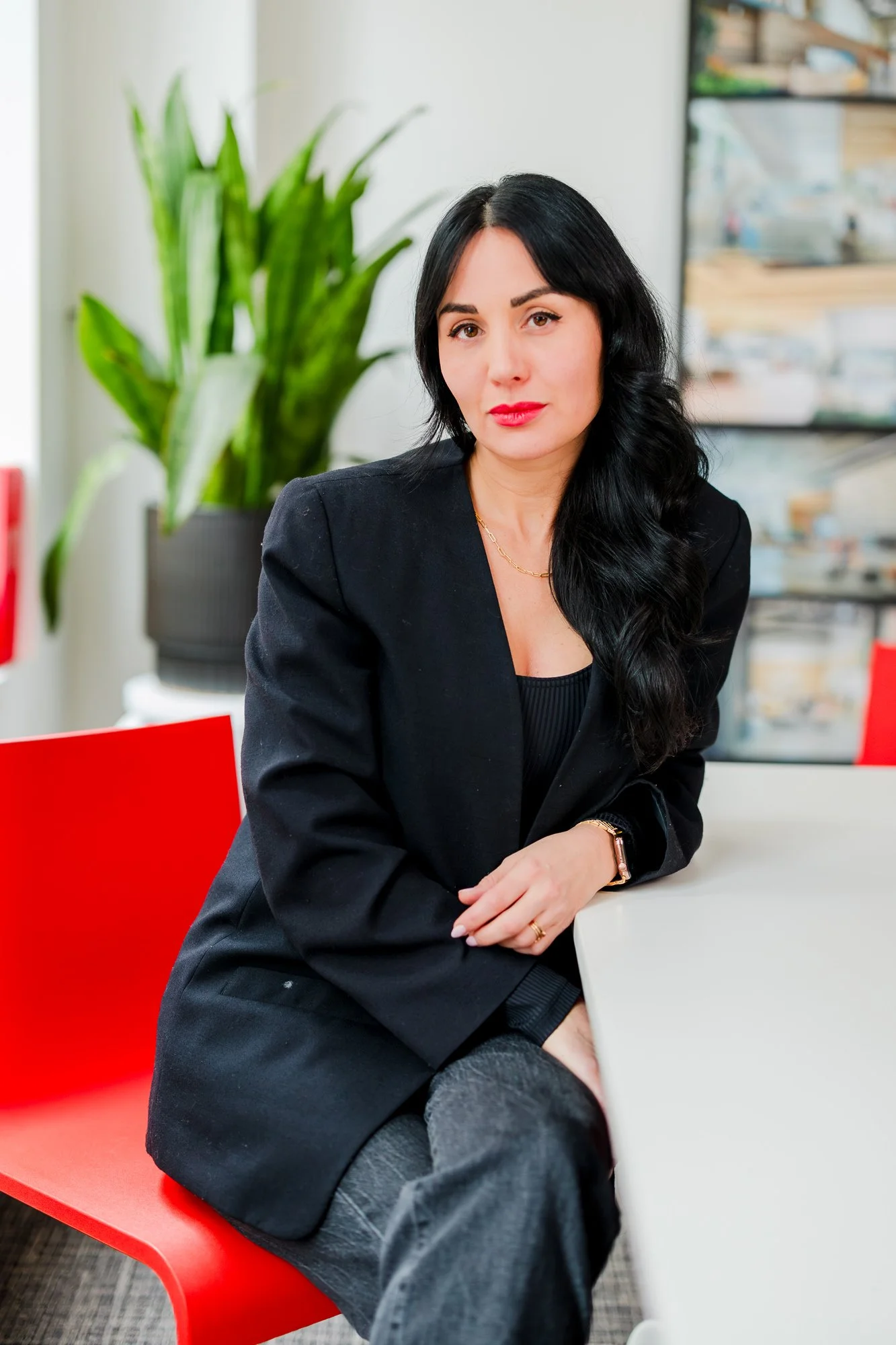 A woman with long black hair and red lipstick sitting at a white table in an office, wearing a black blazer and dark plaid pants, with a green plant and storage boxes in the background.