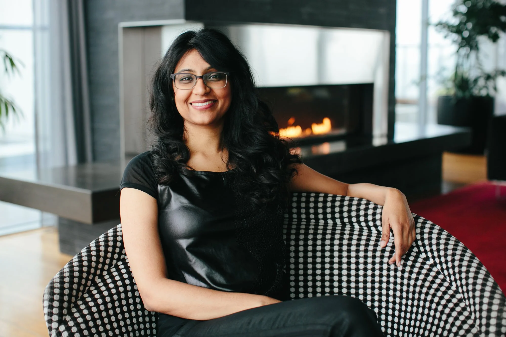 A woman with black curly hair and glasses, smiling and sitting on a patterned chair in a modern living room with a fireplace in the background.