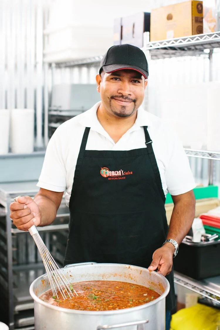 Man in black apron and cap stirring a large pot of red salsa in kitchen