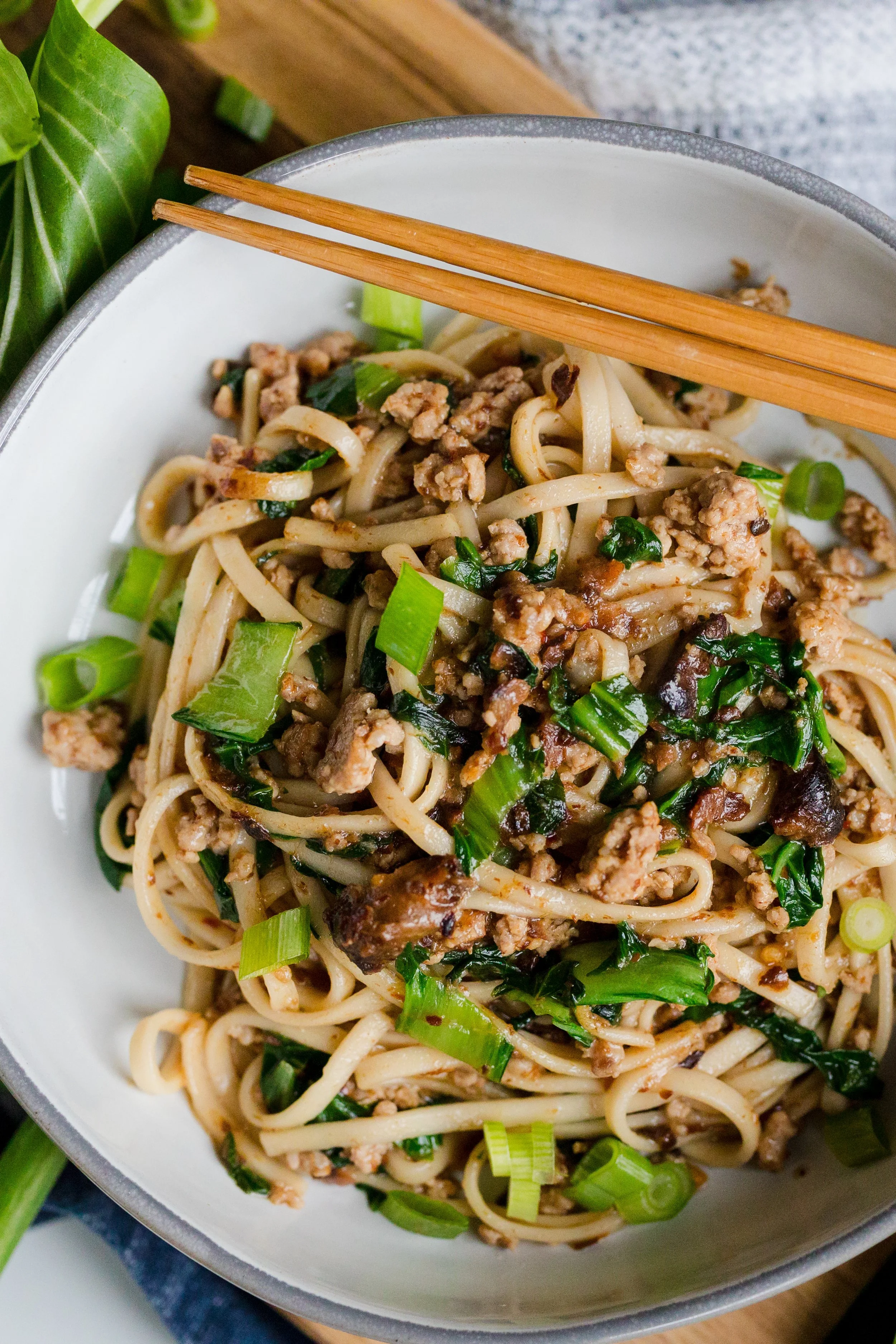 A bowl of stir-fried noodles with ground meat, chopped green onions, and dark leafy greens, with chopsticks resting on the edge.