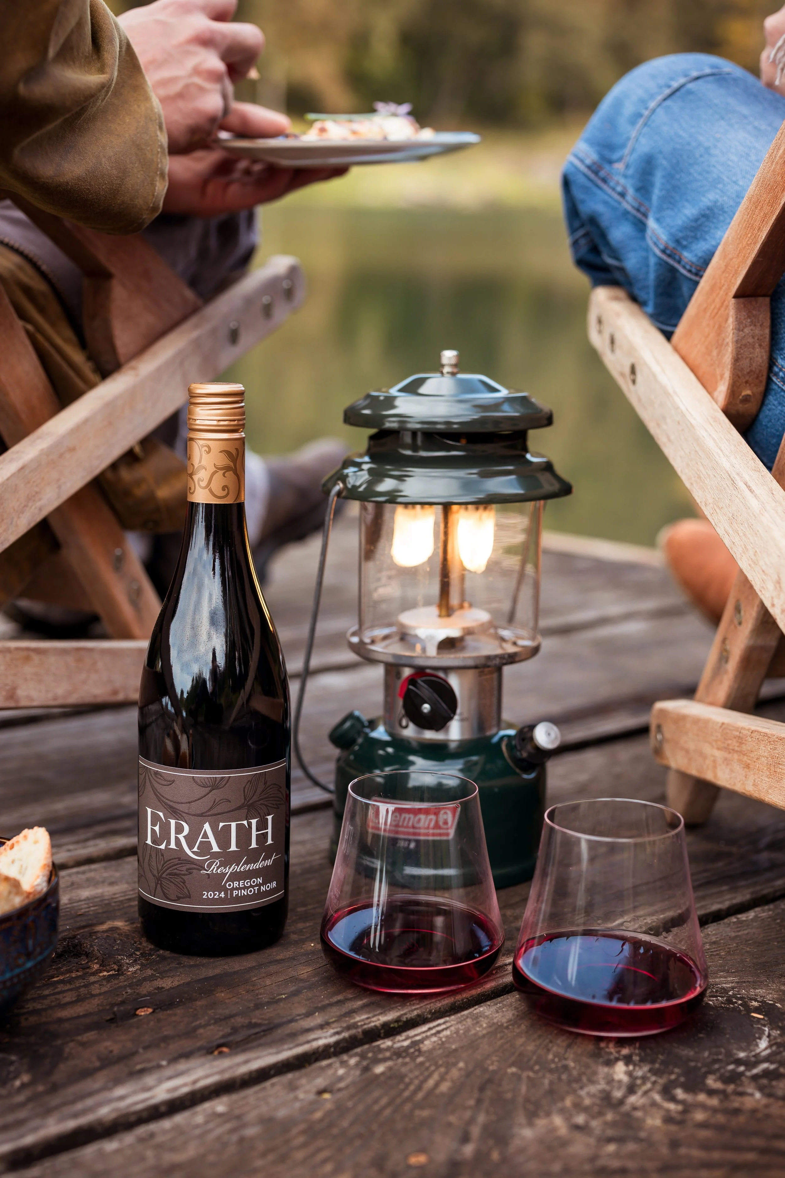 A bottle of red wine, two glasses filled with red wine, and a vintage lantern on a rustic wooden table outdoors near a lake, with people sitting on either side.