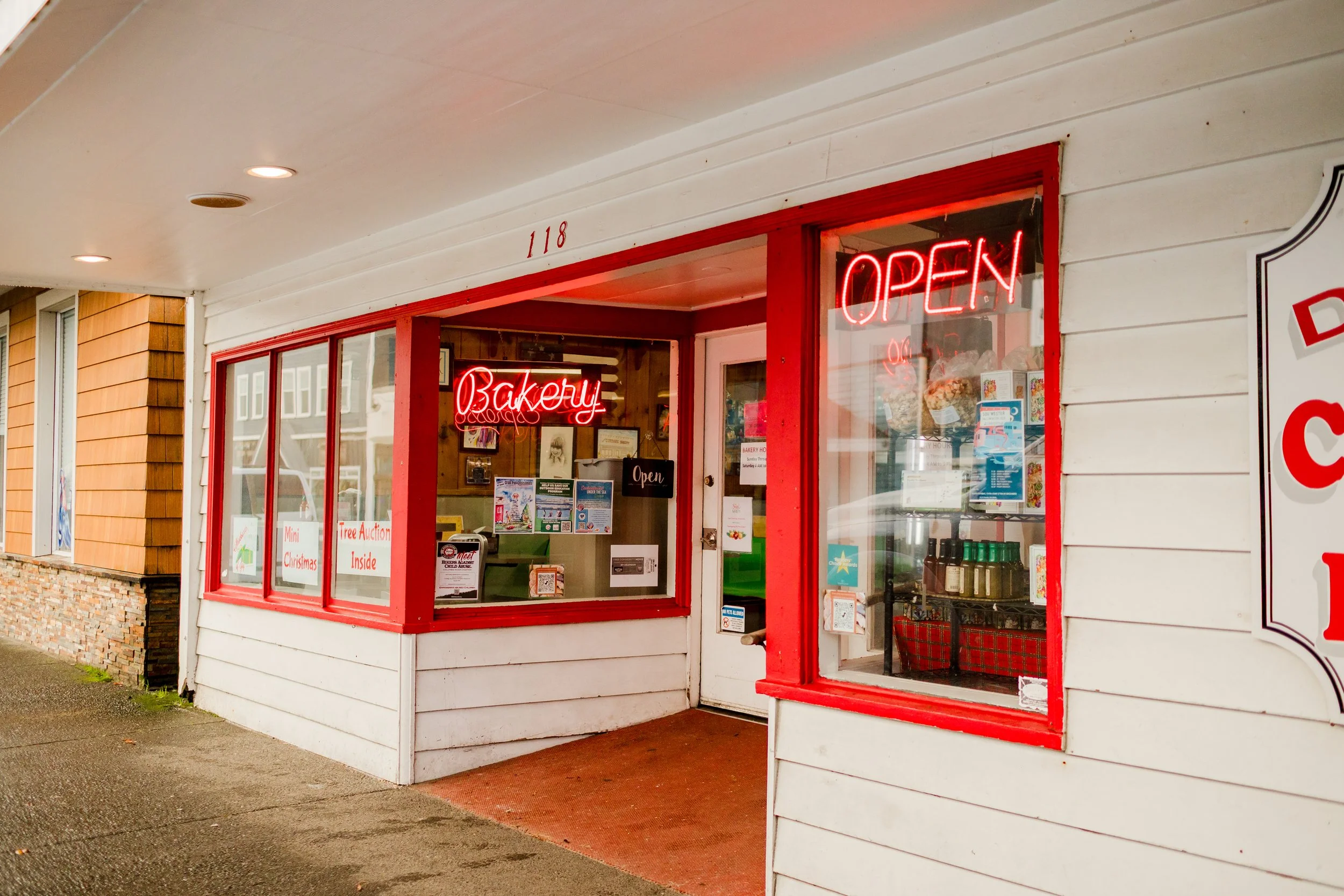 Small bakery storefront with red neon signs reading 'Bakery' and 'Open', display window showing baked goods and spices, and a white exterior with red trim.