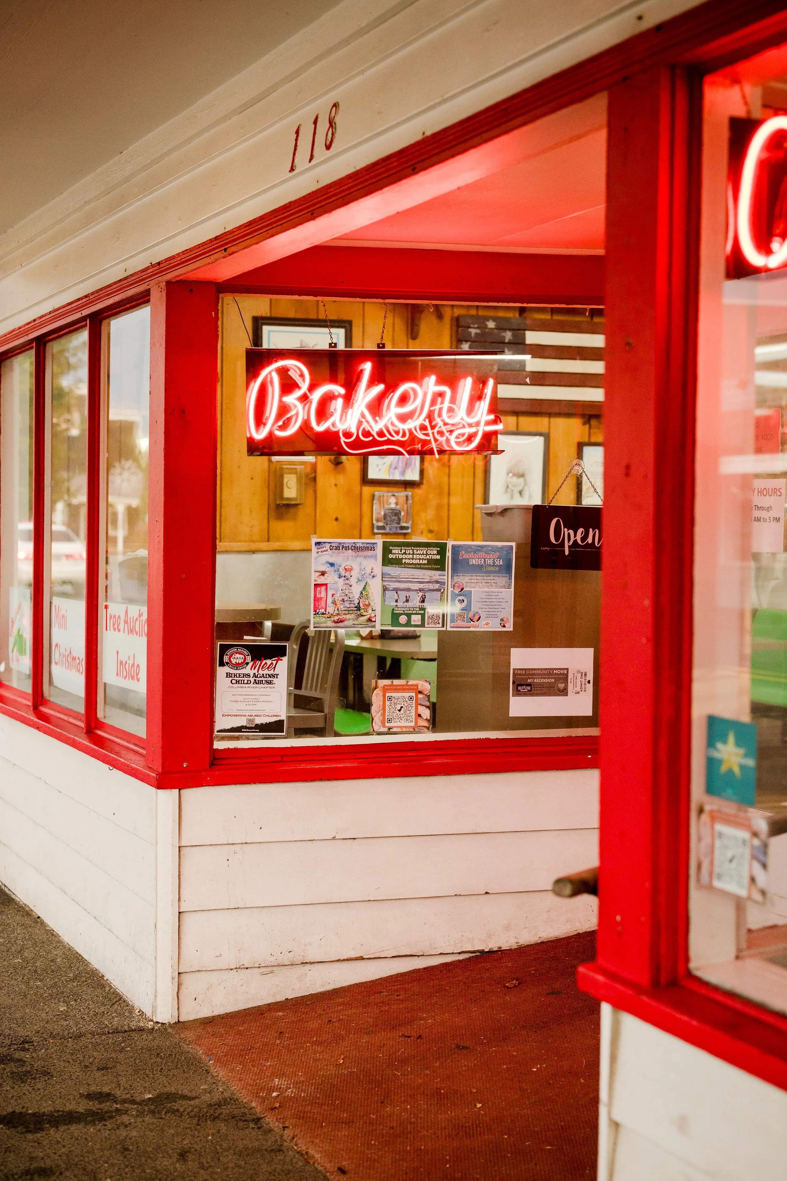 View of a bakery storefront with a neon red 'Bakery' sign, an 'Open' sign, and various posters inside.