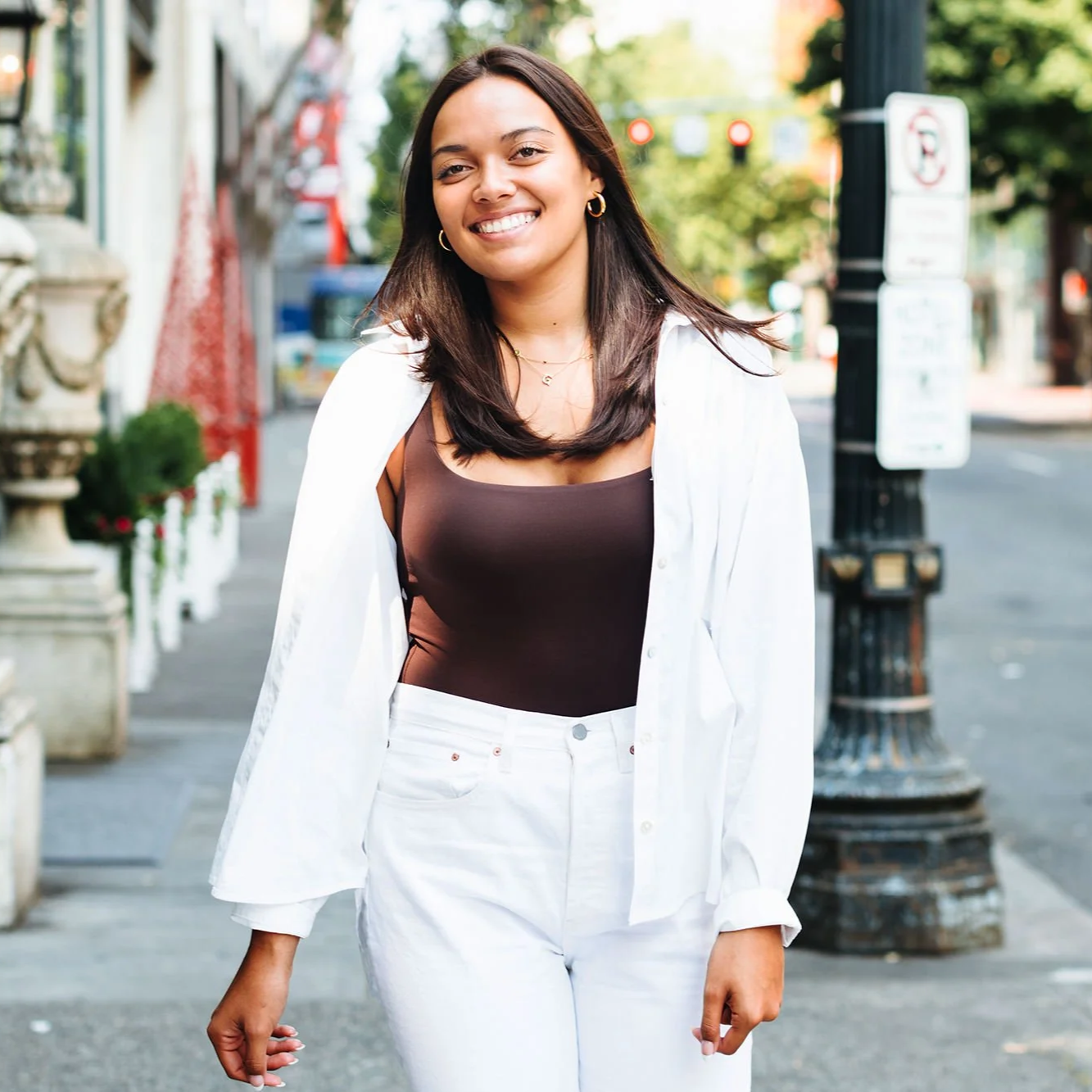 A young woman with dark brown hair smiling and walking on a city sidewalk during daytime. She is wearing a brown sleeveless top, white pants, gold hoop earrings, and a white shirt draped over her shoulders.