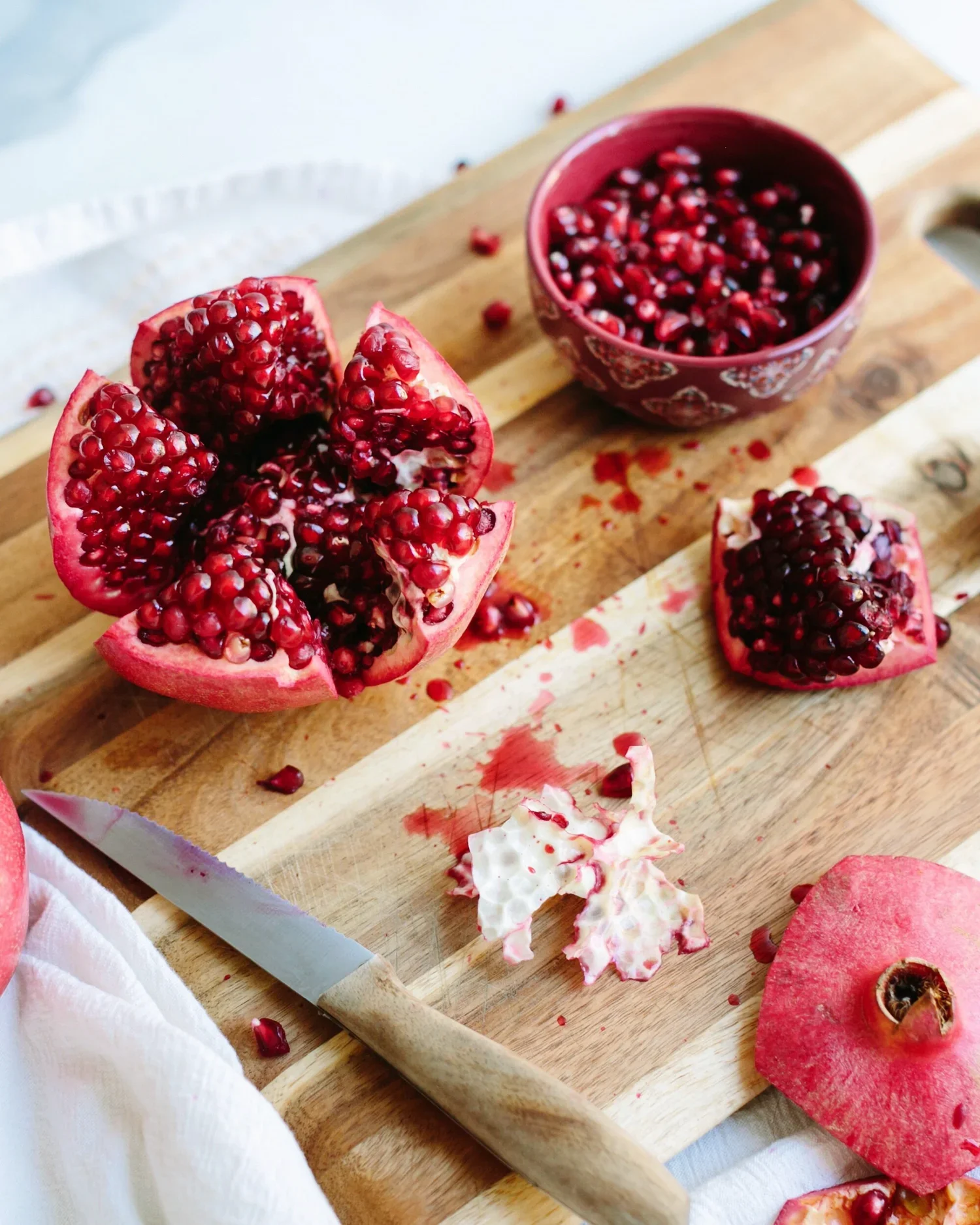 Pomegranate with opened seeds, a small bowl of pomegranate seeds, and a partially peeled pomegranate on a wooden cutting board with a knife.