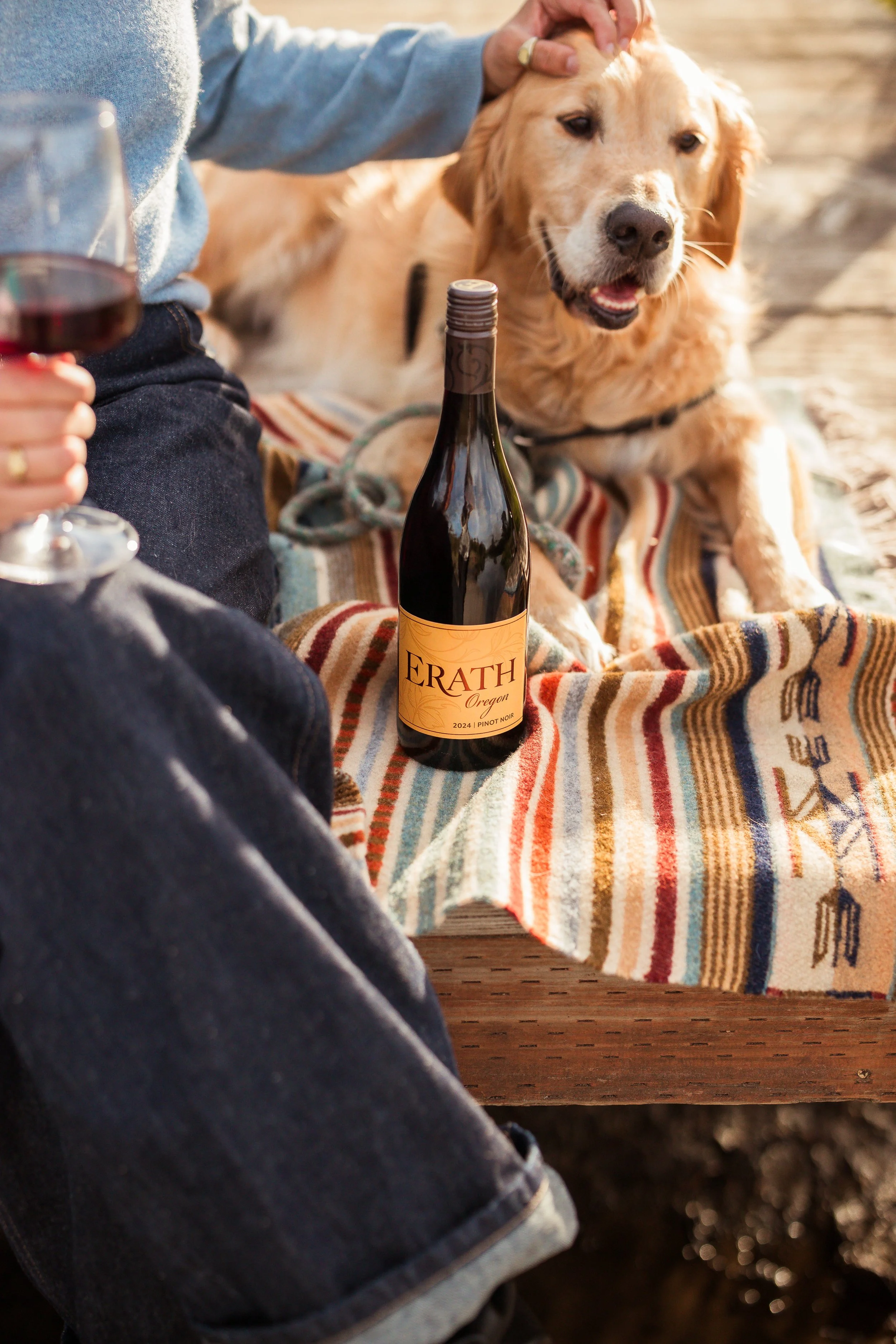 Person holding a glass of red wine with a dog lying on a blanket beside a bottle of Erath Oregon Pinot Noir wine at an outdoor setting.