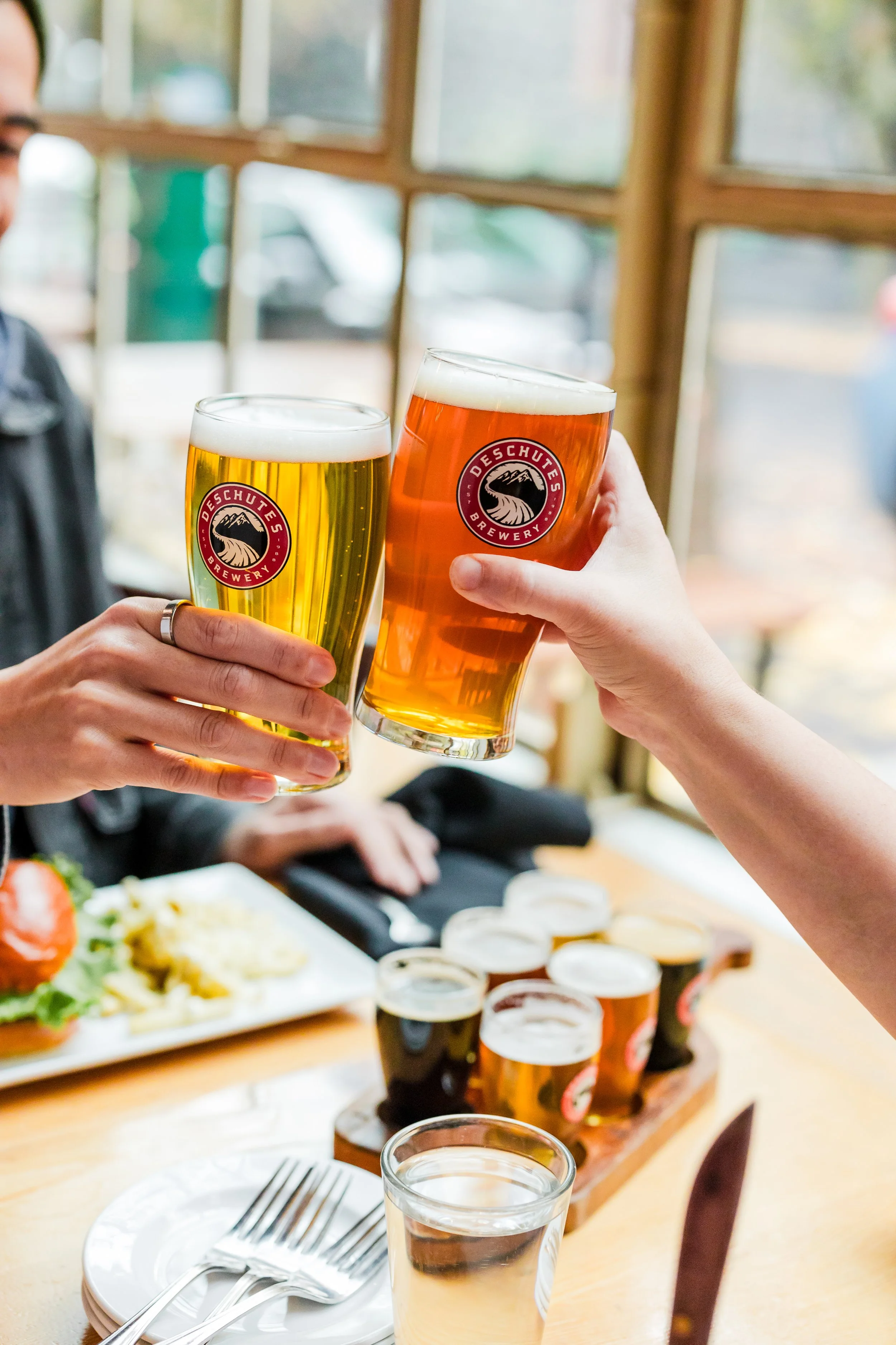 Two people toasting with brewery glasses of beer, with additional beer glasses on a table and food in the background.